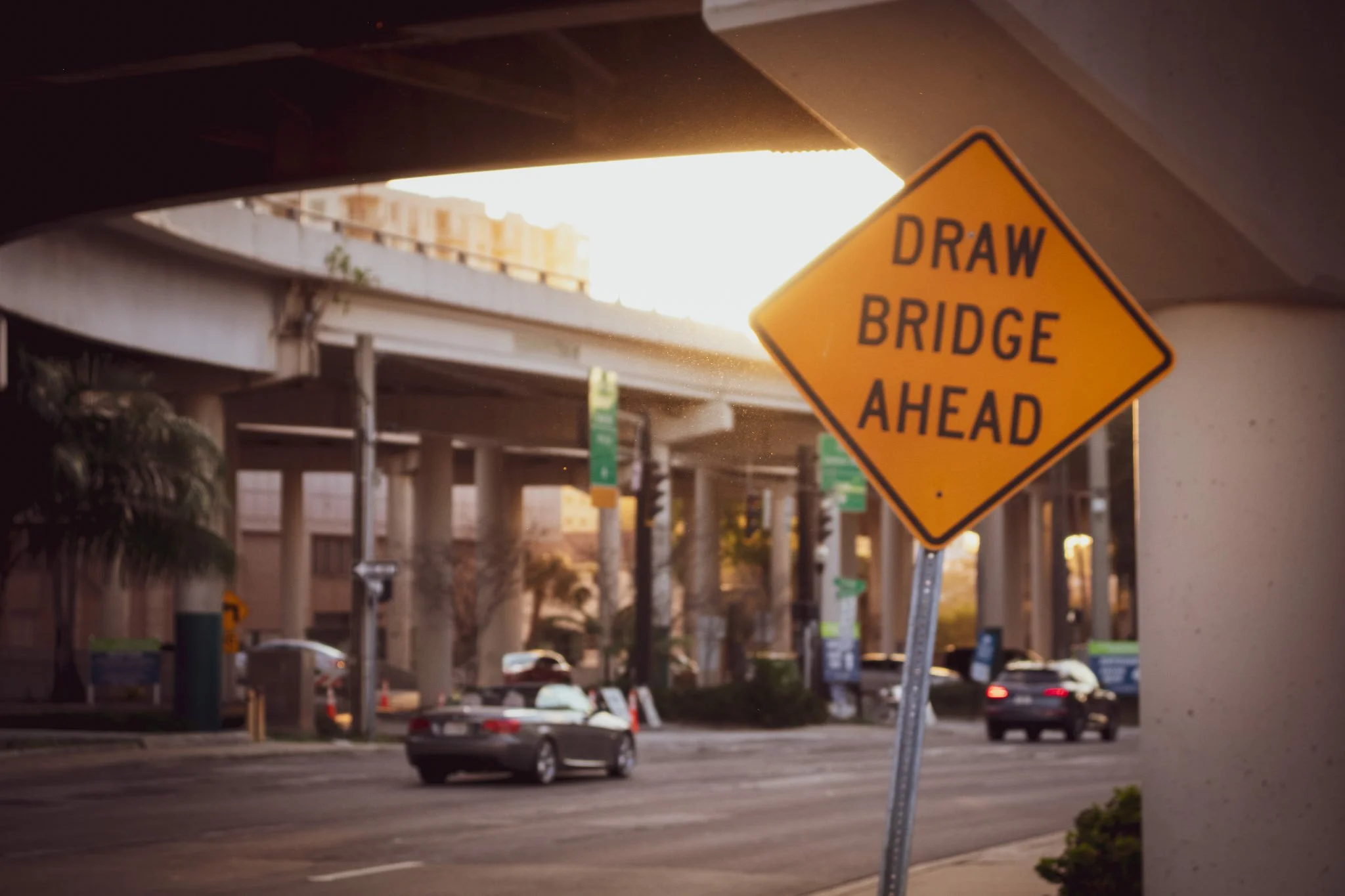 A yellow diamond-shaped road sign that reads 'Draw Bridge Ahead' is mounted on a pole beneath a highway overpass. The overpass and surrounding cityscape are visible in the background, with cars on the road and a sunset lighting the scene.
