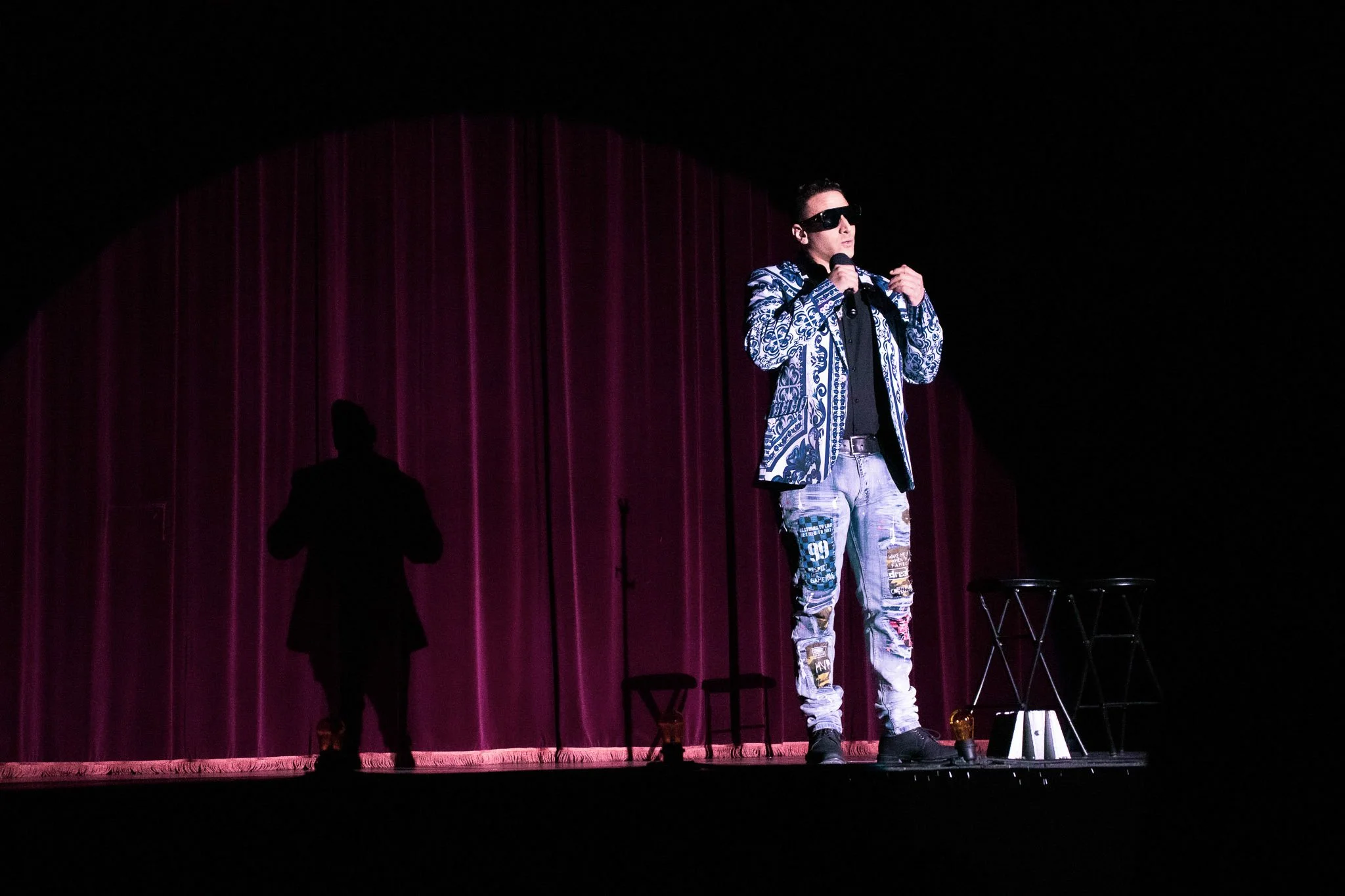 Person on stage wearing sunglasses, patterned jacket, and ripped jeans, holding a microphone in front of a purple curtain.