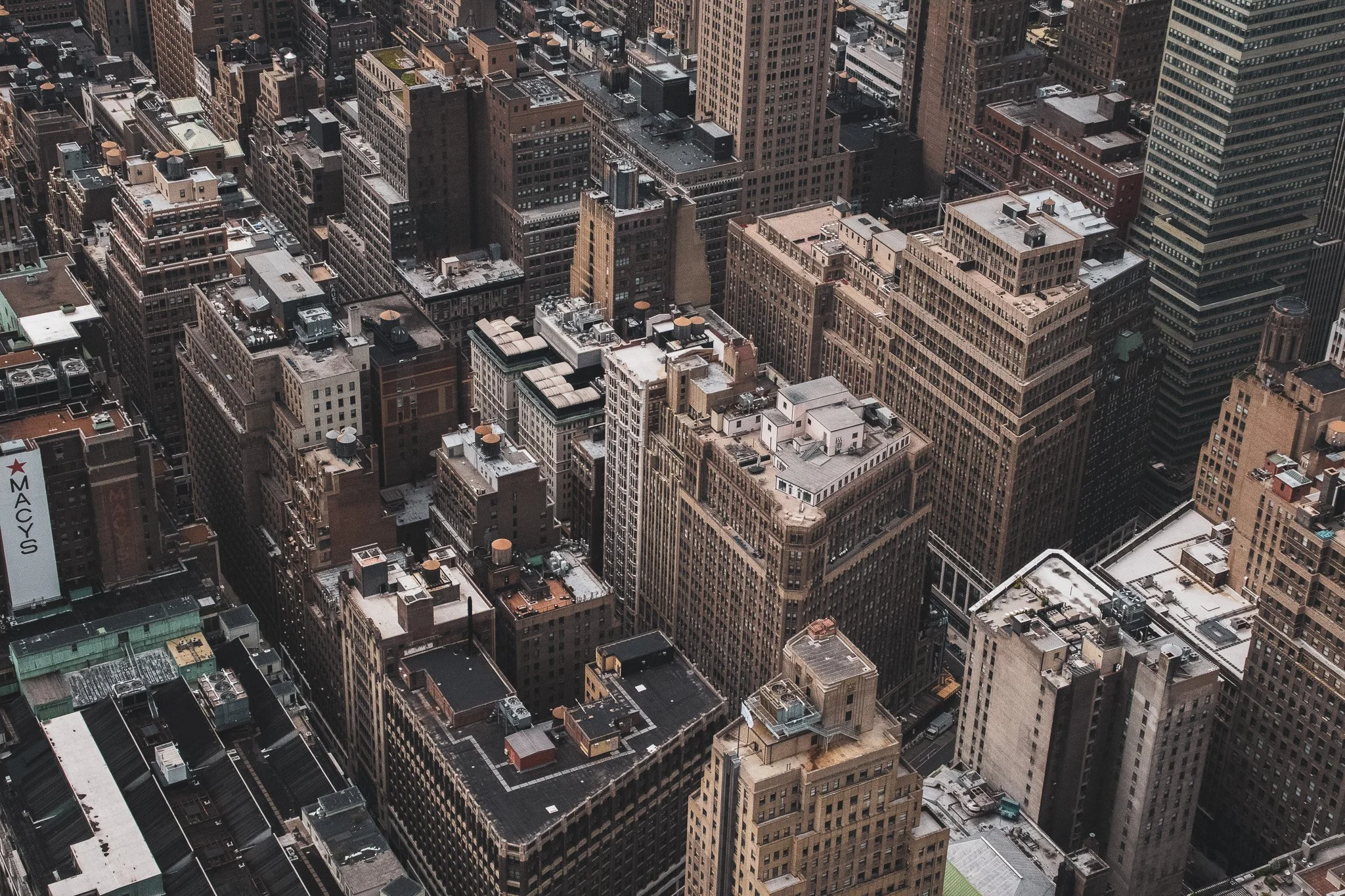 Aerial view of a dense cityscape with numerous tall skyscrapers and buildings of varying heights and architectural styles, some with rooftop equipment and water tanks.