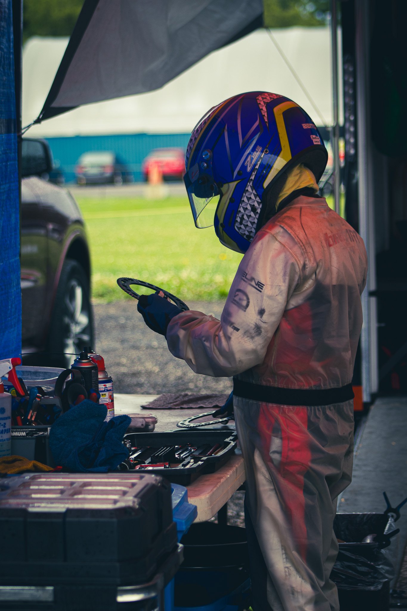 A person wearing a colorful helmet and a racing suit standing at a workbench with tools and supplies, preparing for motorsport activities.
