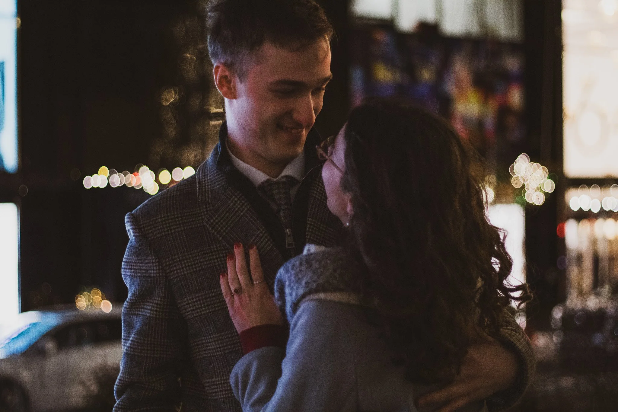 A young man in a checkered suit and a young woman in a sweater are sharing a close moment, possibly a dance, in a dimly lit setting with blurred colorful lights in the background.