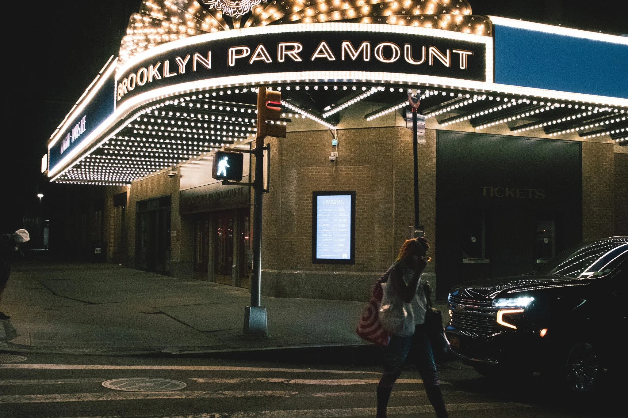 Nighttime view of the Brooklyn Paramount Theater marquee with bright lights and the sign reading 'Brooklyn Paramount'. Pedestrians and cars are visible on the street.