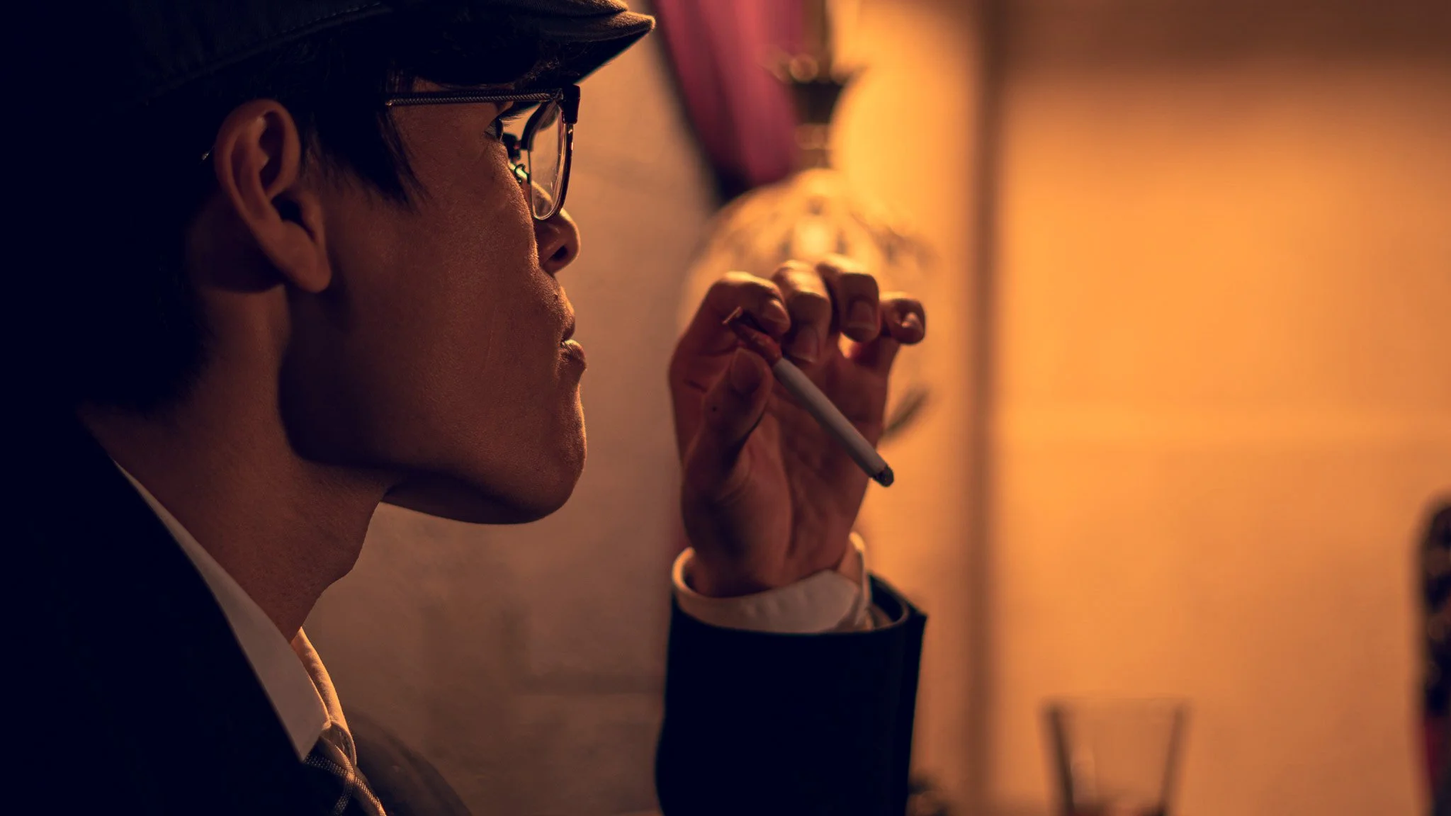 Side profile of a man wearing glasses and a hat, holding a cigarette near his lips in a dimly lit room.