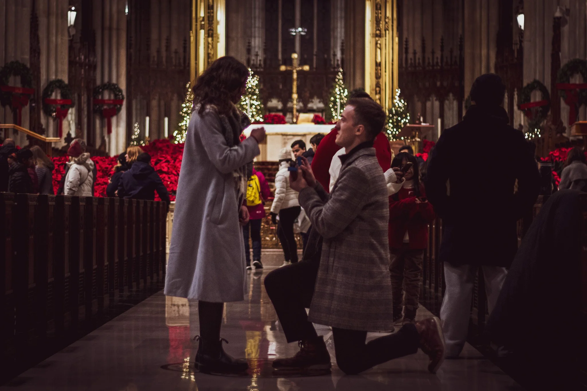 A man on one knee proposing to a woman inside a church decorated for Christmas, with other people gathered around.