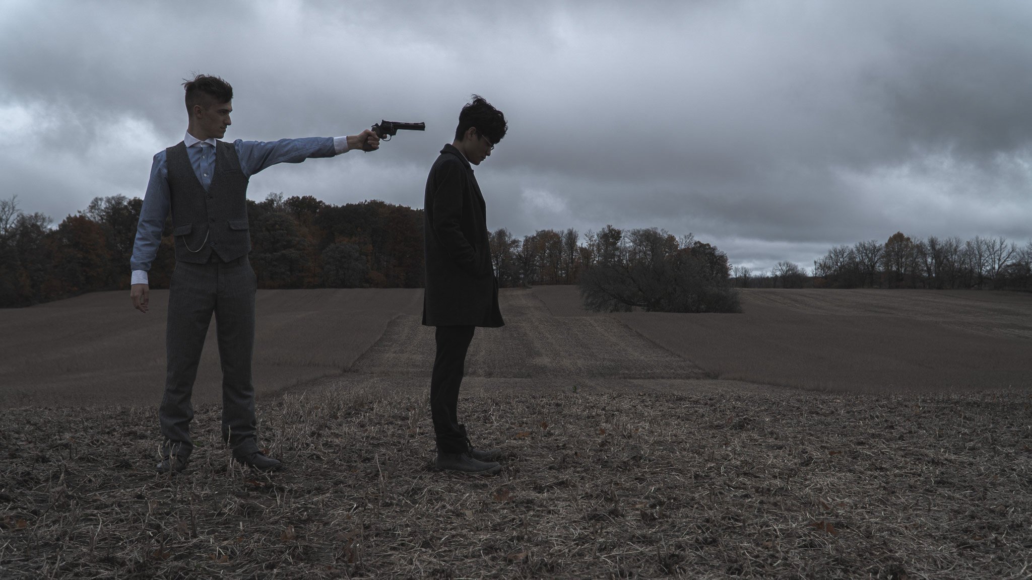Two young men outdoors on a cloudy day; one is pointing a gun at the other's head, and the other is looking down, appearing resigned.