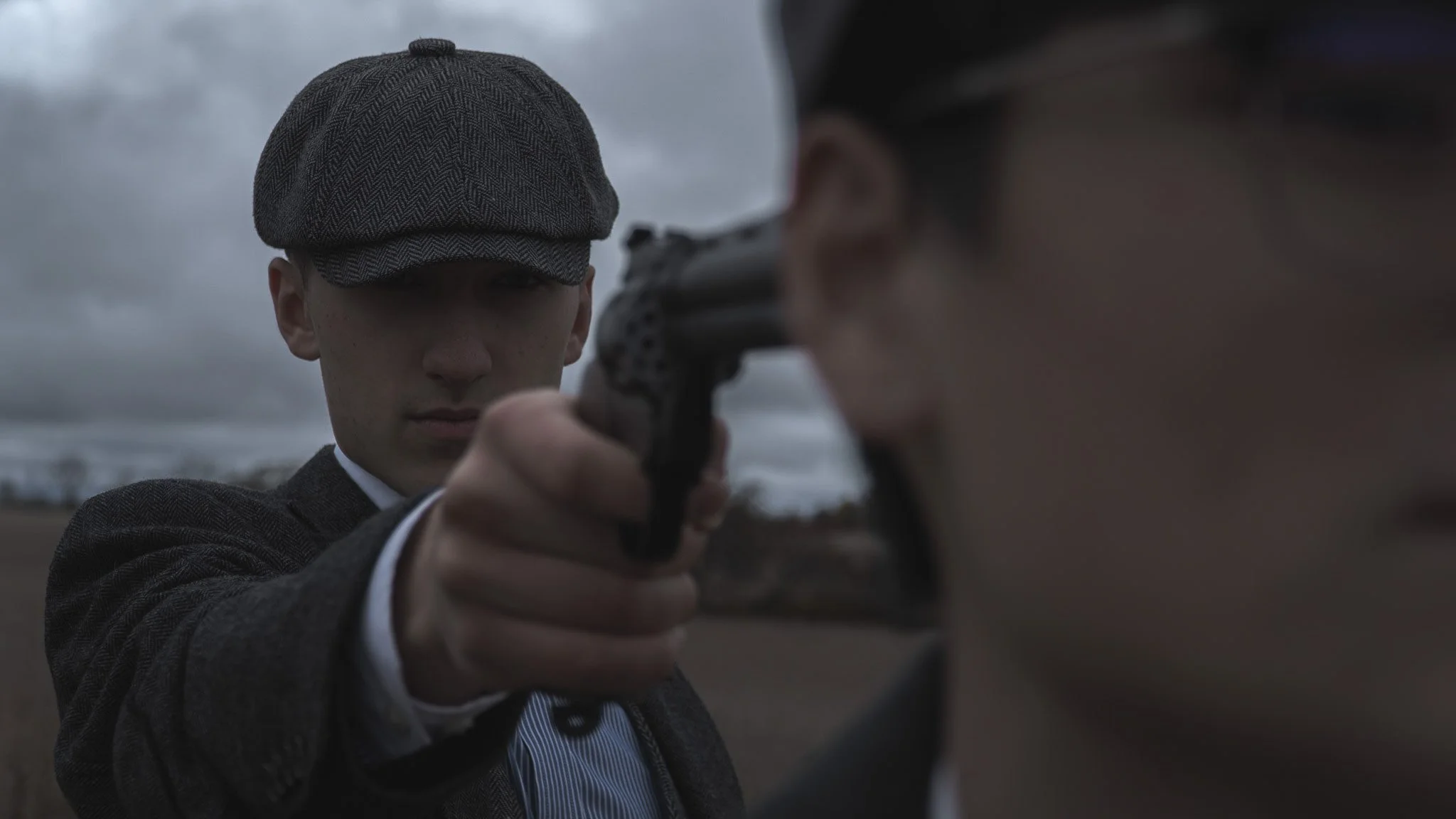 A person wearing a flat cap and a herringbone suit pointing a pistol at the camera, with an overcast sky in the background.