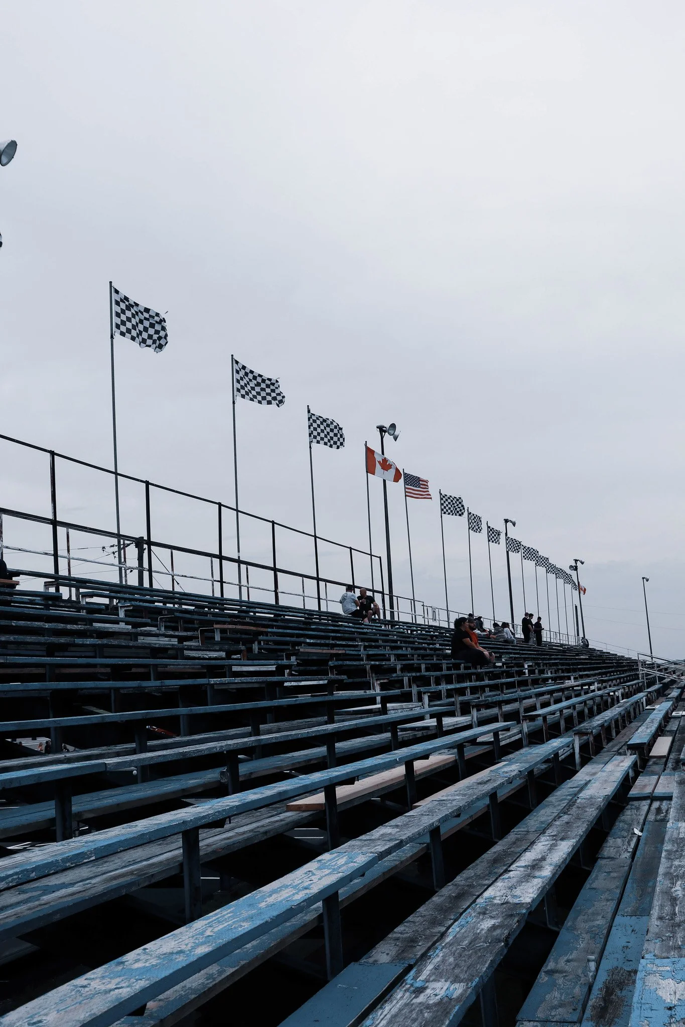 View of empty bleachers at a race track with several flags, including checkered flags, the Canadian flag, and the American flag, against a cloudy sky.