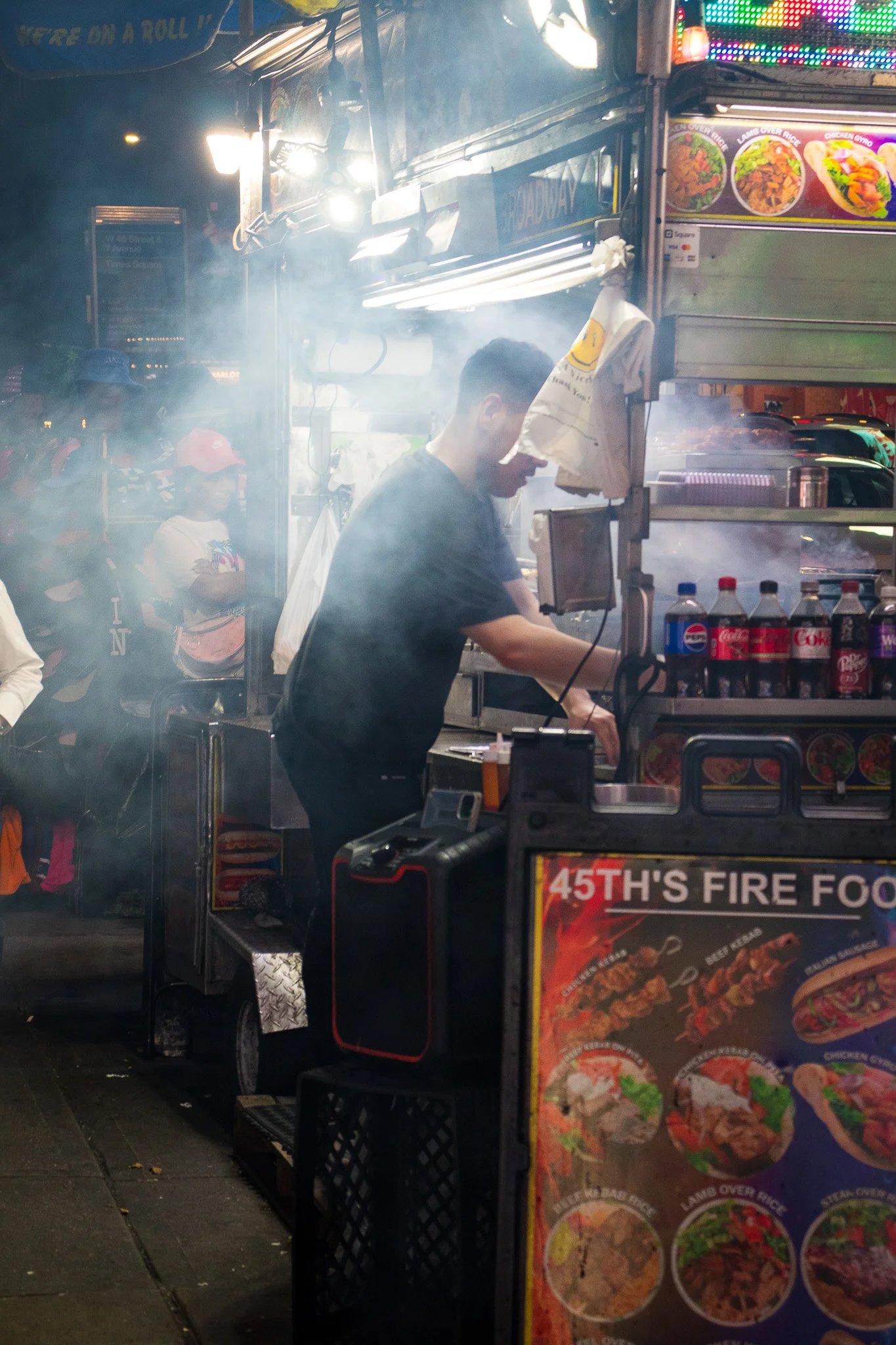 A street food stall at night with a man preparing food, illuminated by bright lights and surrounded by smoke. Children wearing masks and a woman are visible in the background. The stall displays colorful menus with pictures of various dishes.