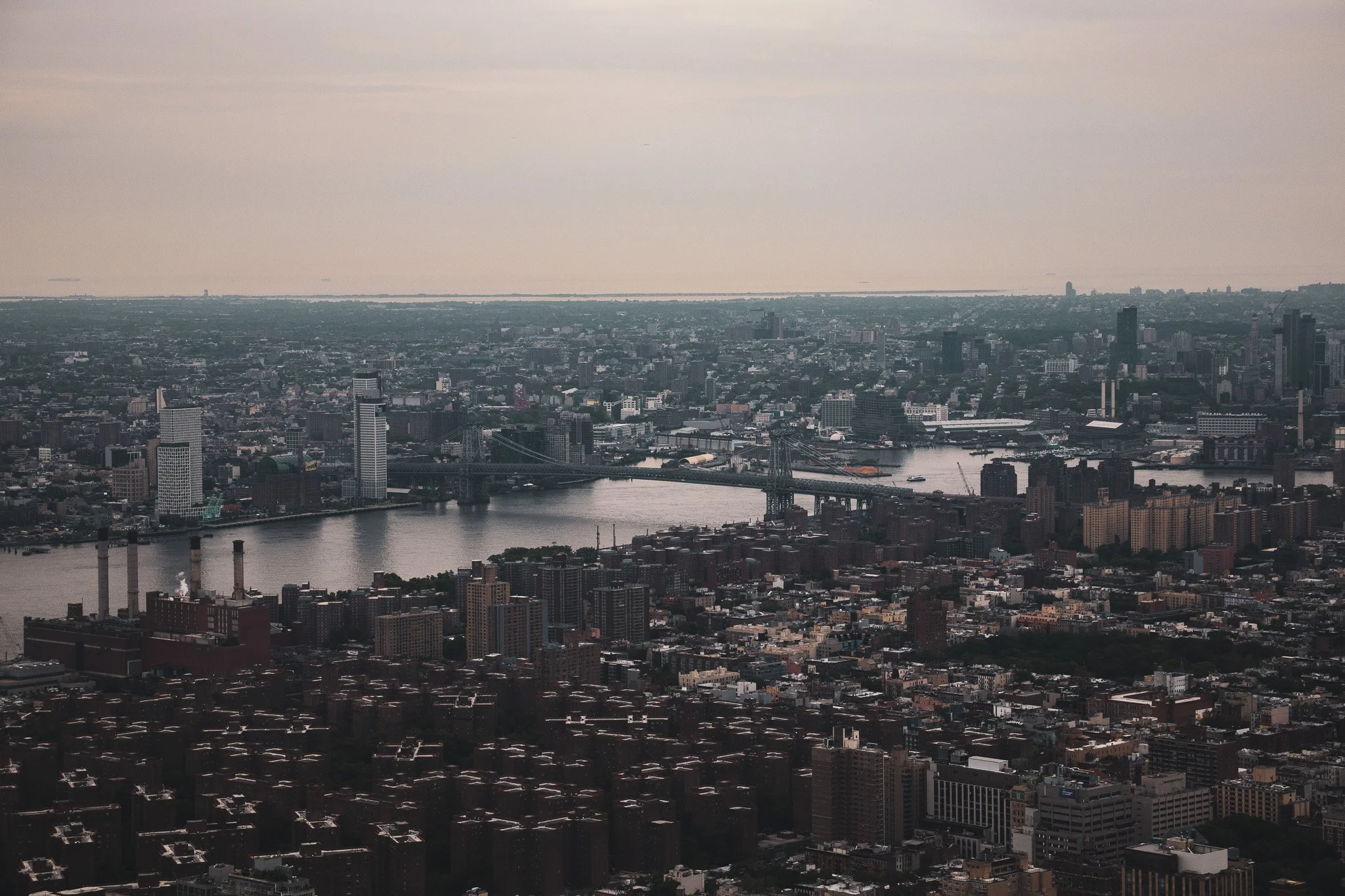 A panoramic view of a cityscape showing numerous buildings, a river with a bridge, and a distant horizon with water and islands, under a cloudy sky.
