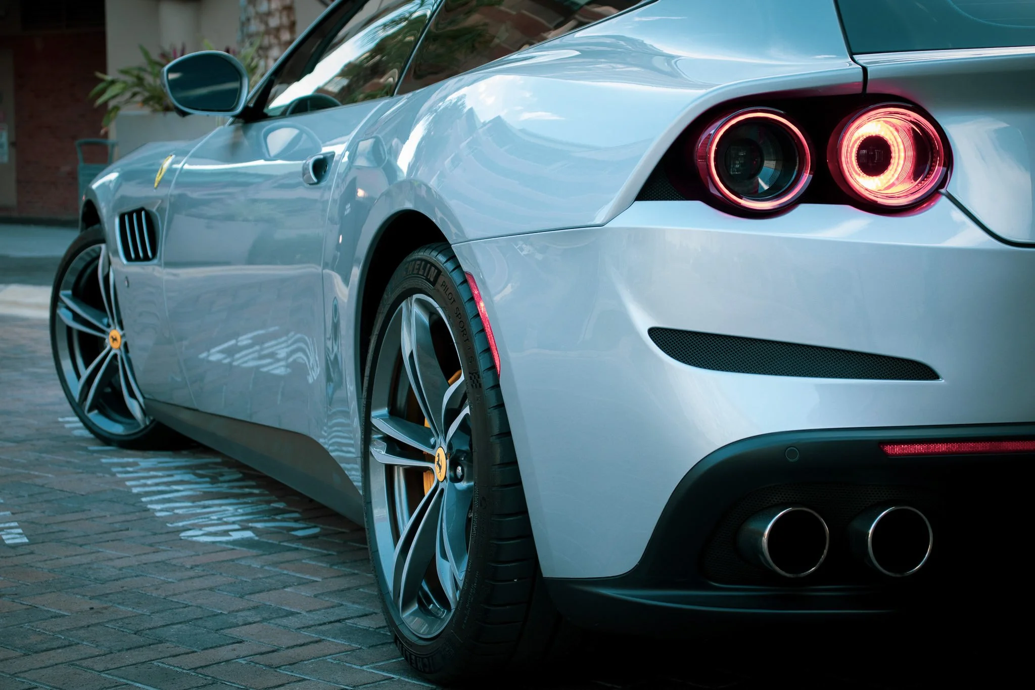 Close-up of a silver sports car parked on a brick street, focusing on the rear right side, showing the taillights, wheel, and exhaust pipes.