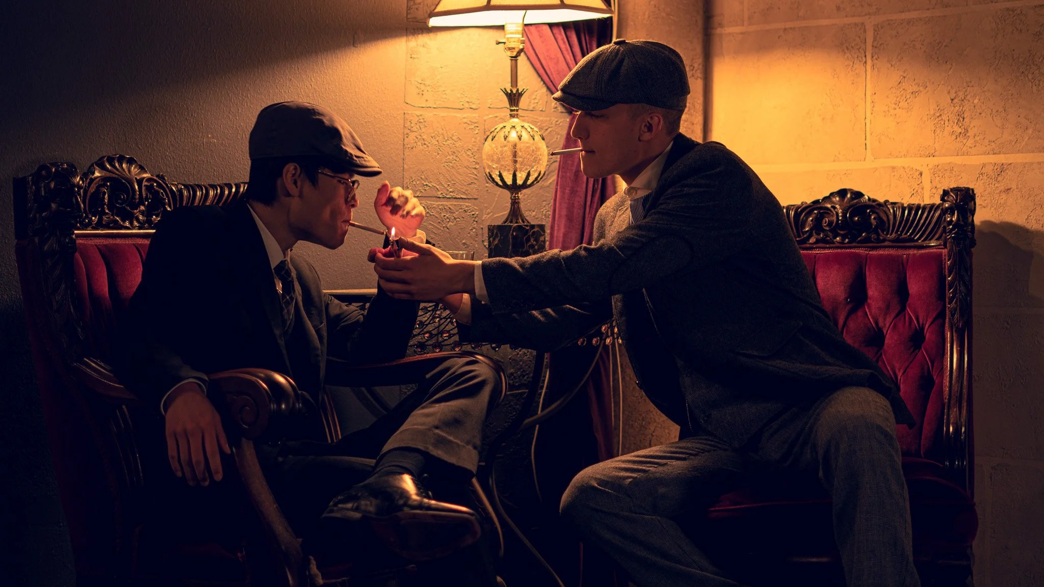 Two men in vintage clothing, wearing flat caps and glasses, engage in a cigarette lighting exchange in a dimly lit room with ornate furniture and a table lamp.
