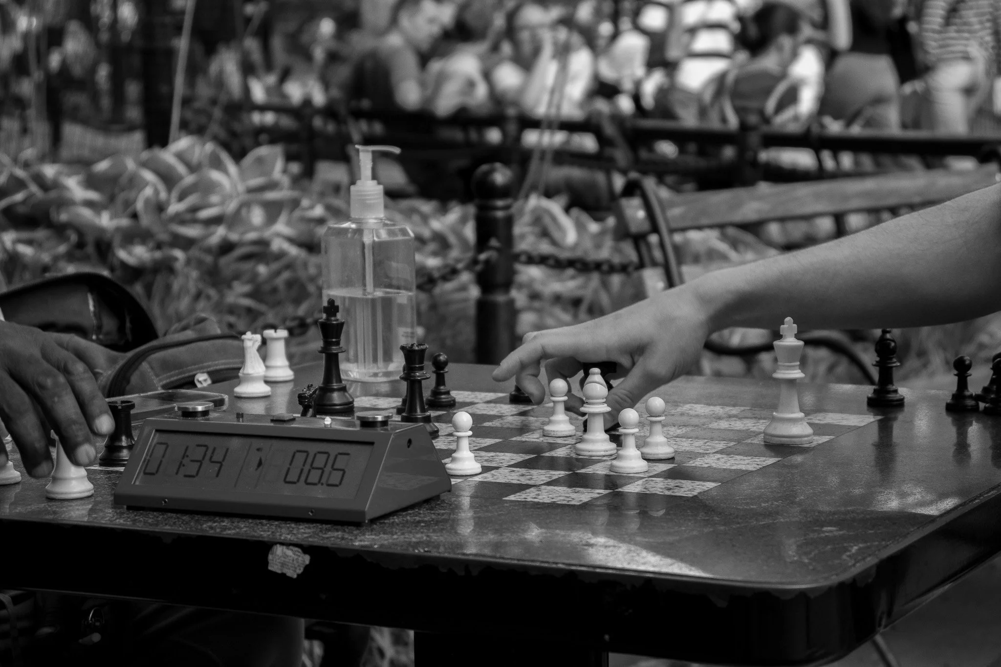 Two people playing chess at a table with a chess clock and a water bottle in the background, in a busy indoor setting.