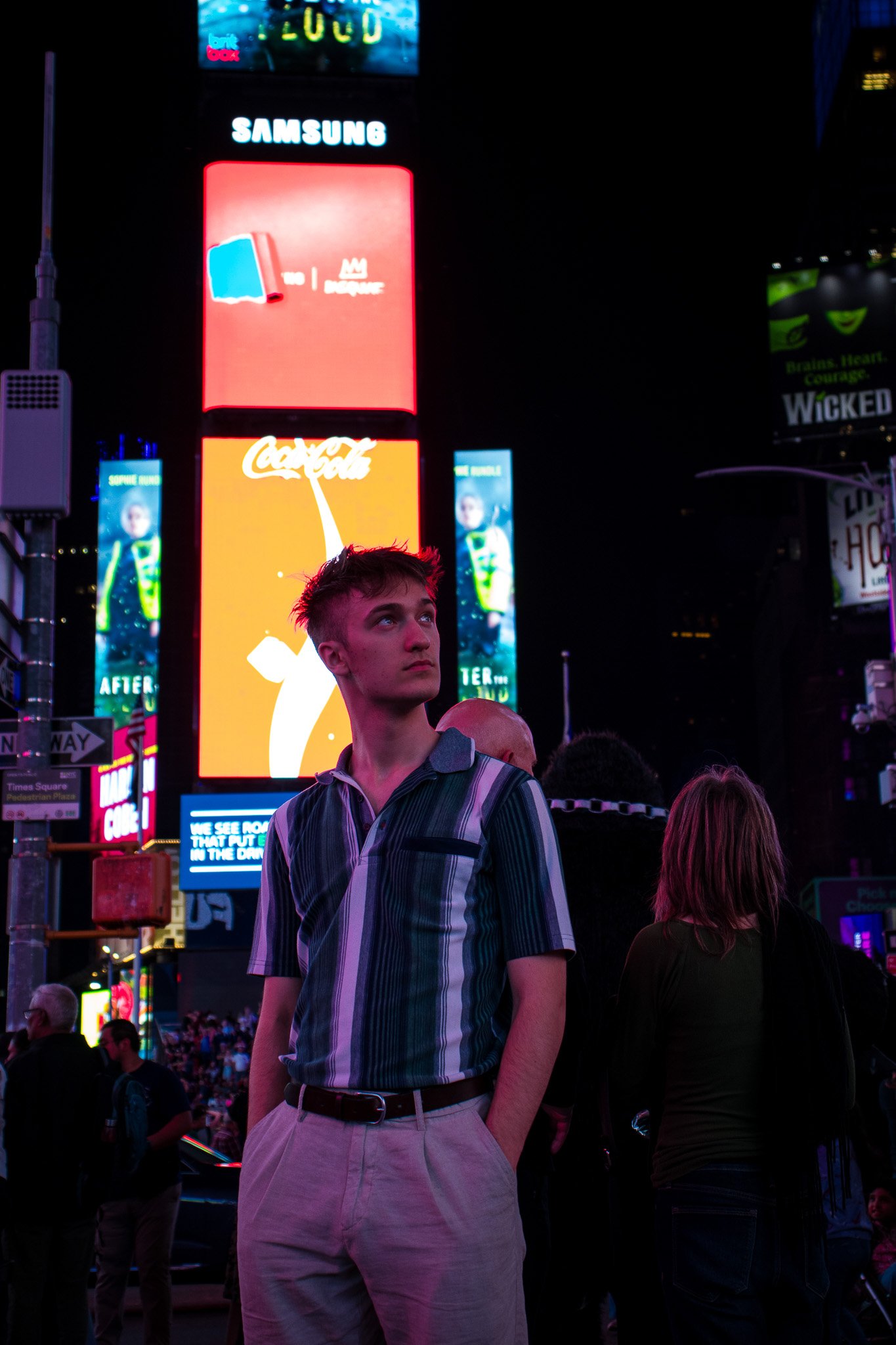 Nighttime scene with digital billboards and neon signs in Times Square. A young man with short hair, wearing a striped shirt, stands in the foreground looking to the side. Other people are visible around him, some facing away. Bright lights and adver