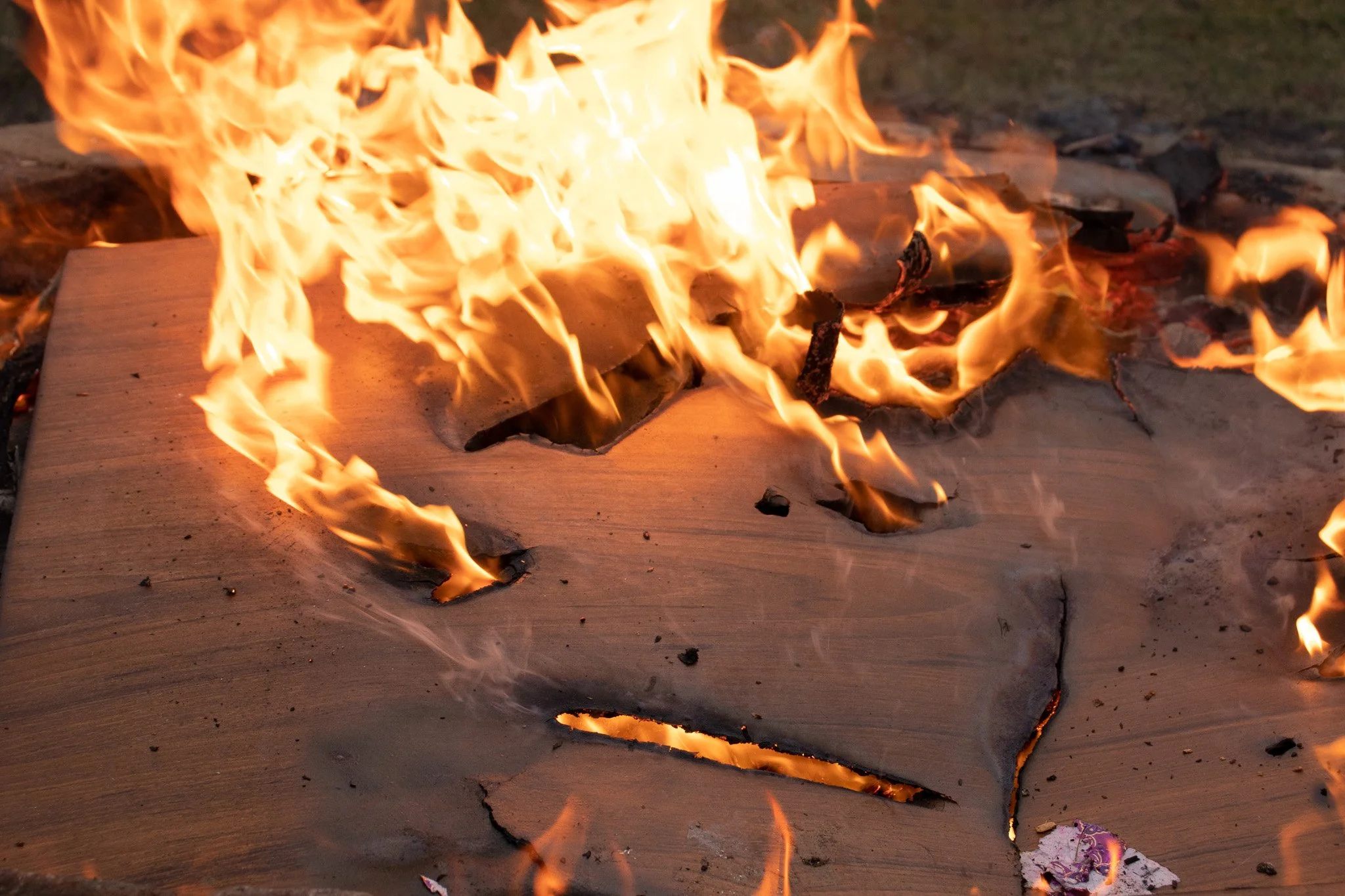 An open fire burning on a wooden surface outdoors with visible flames and burnt debris.