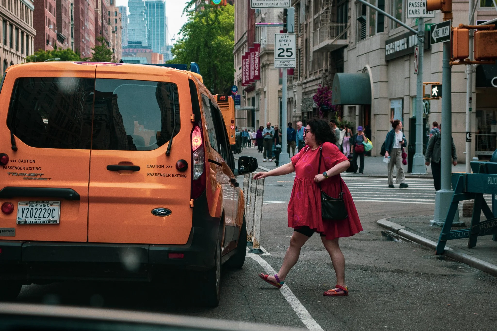 A woman wearing a red dress and sandals is crossing the street, holding a small bag, next to an orange taxi cab in an urban cityscape. There are people walking along the sidewalk, street signs, and tall buildings.