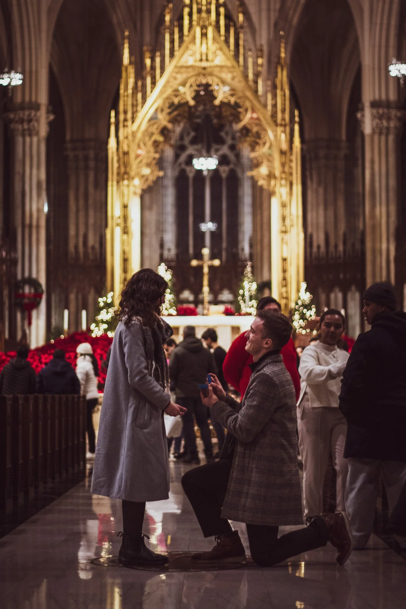 A man proposes marriage to a woman inside a church decorated for Christmas, with people observing and Christmas trees with lights in the background.