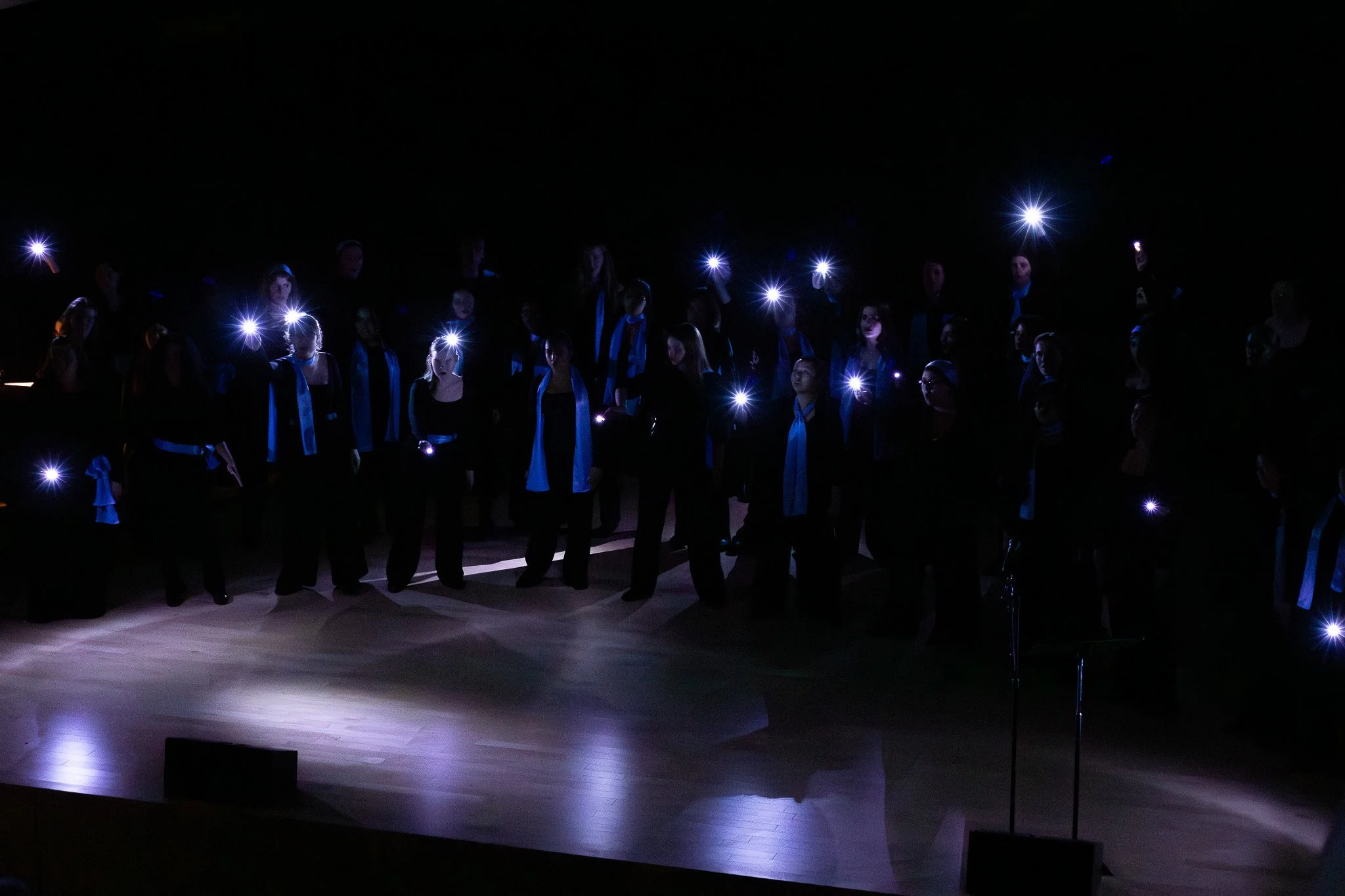Group of people wearing formal attire standing on stage in a dark theater, illuminated by small lights from handheld devices or flashlights.