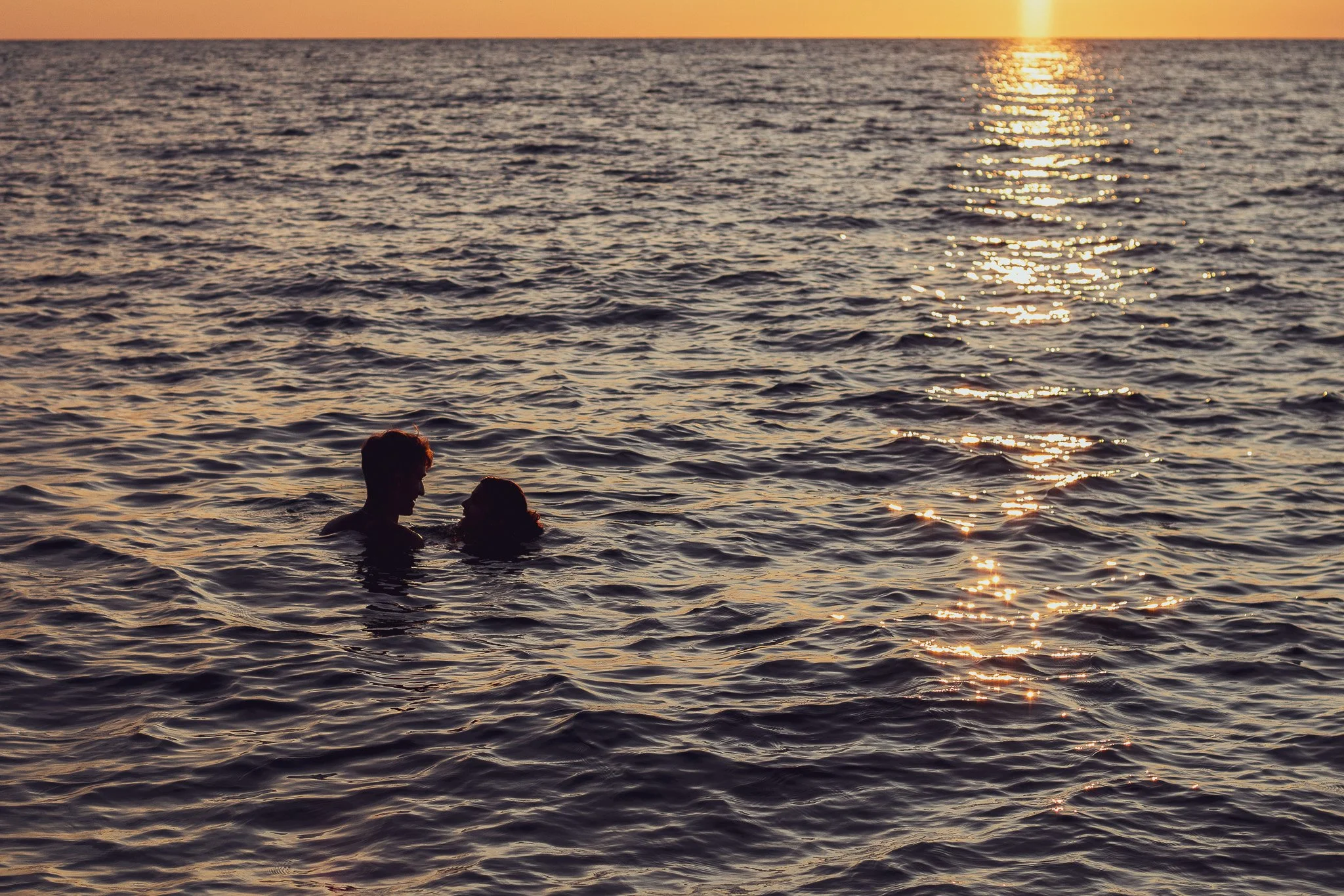 Two people swimming in the ocean during sunset, with the sun reflecting on the water.