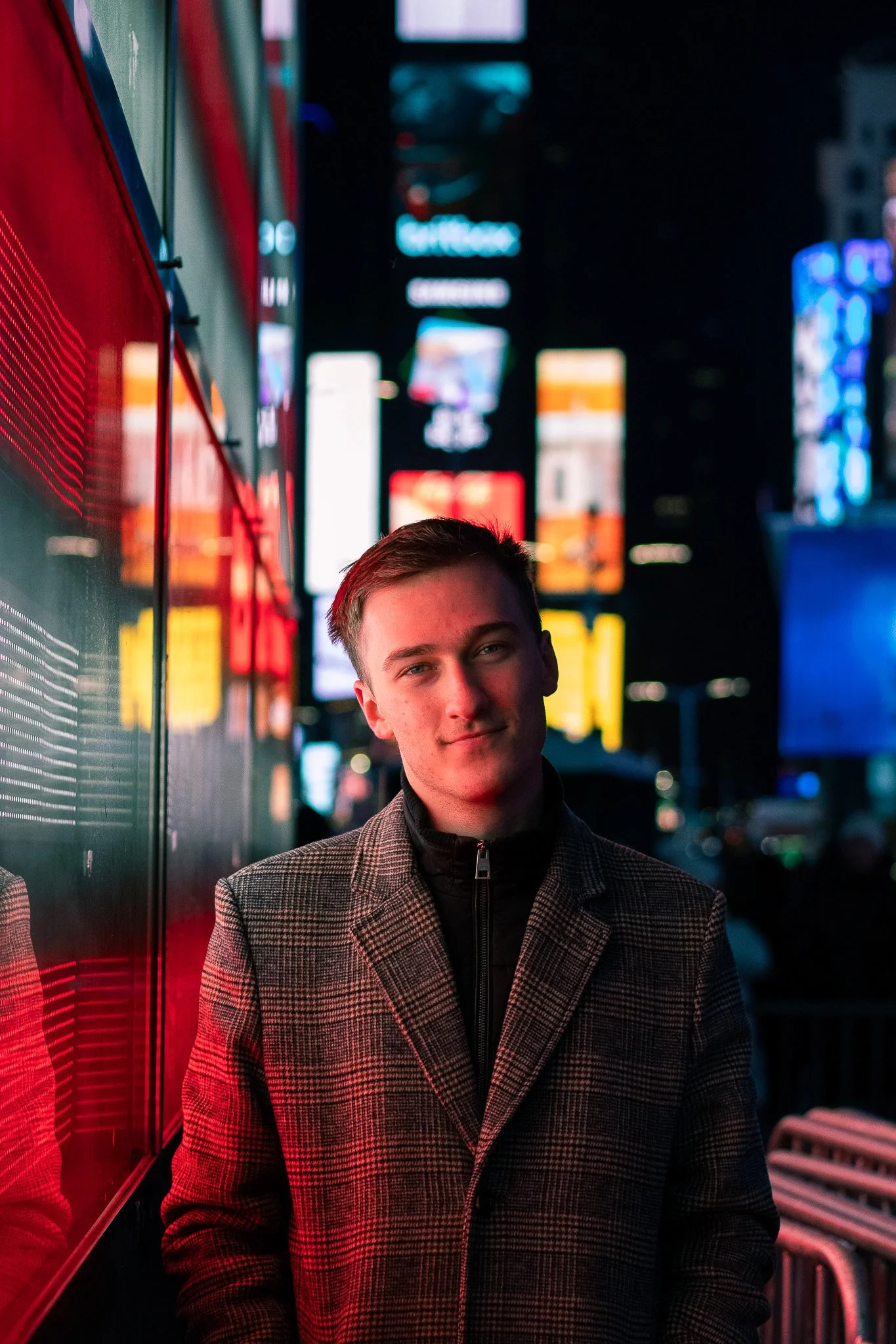 A young man standing at night under neon lights in a city. He is wearing a checkered coat and black jacket, smiling softly. The background features colorful illuminated signs and blurred cityscape.