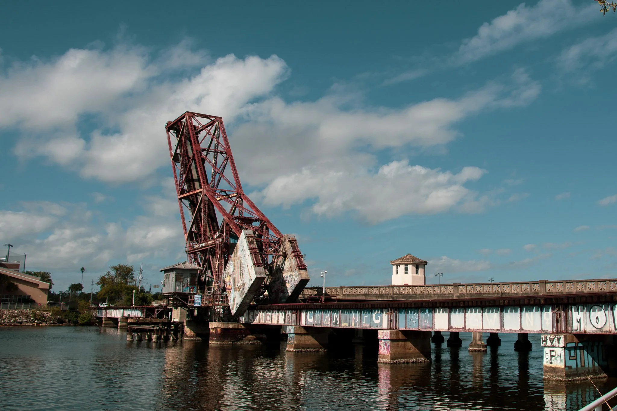An old, rusted amusement park ride structure on a pier over water, with a small building nearby, under a cloudy sky.