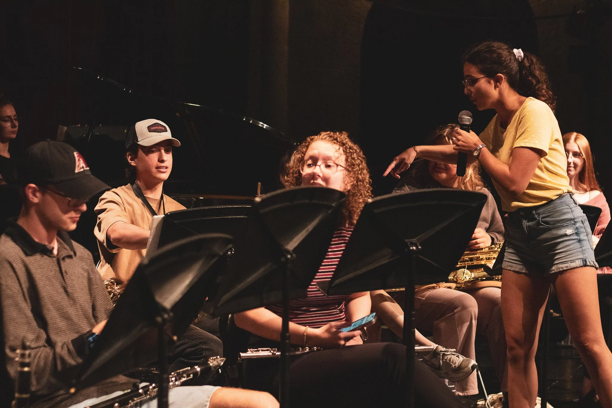 A woman in a yellow shirt and glasses holding a microphone, pointing at a seated woman with curly hair and glasses during a musical rehearsal or performance.