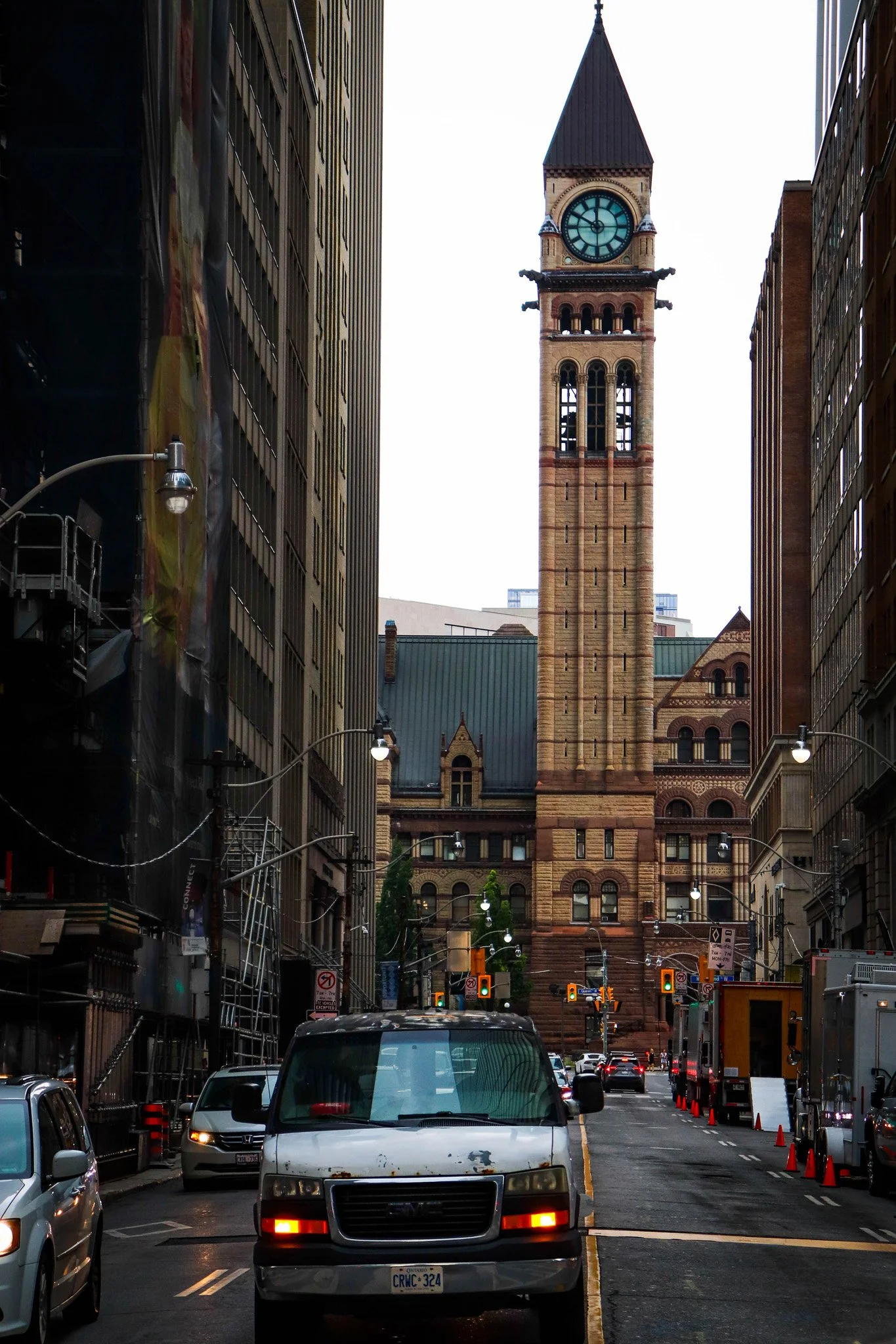 Street view in downtown with a clock tower in the background, lined with tall buildings, cars on the road, and construction cones along the side.
