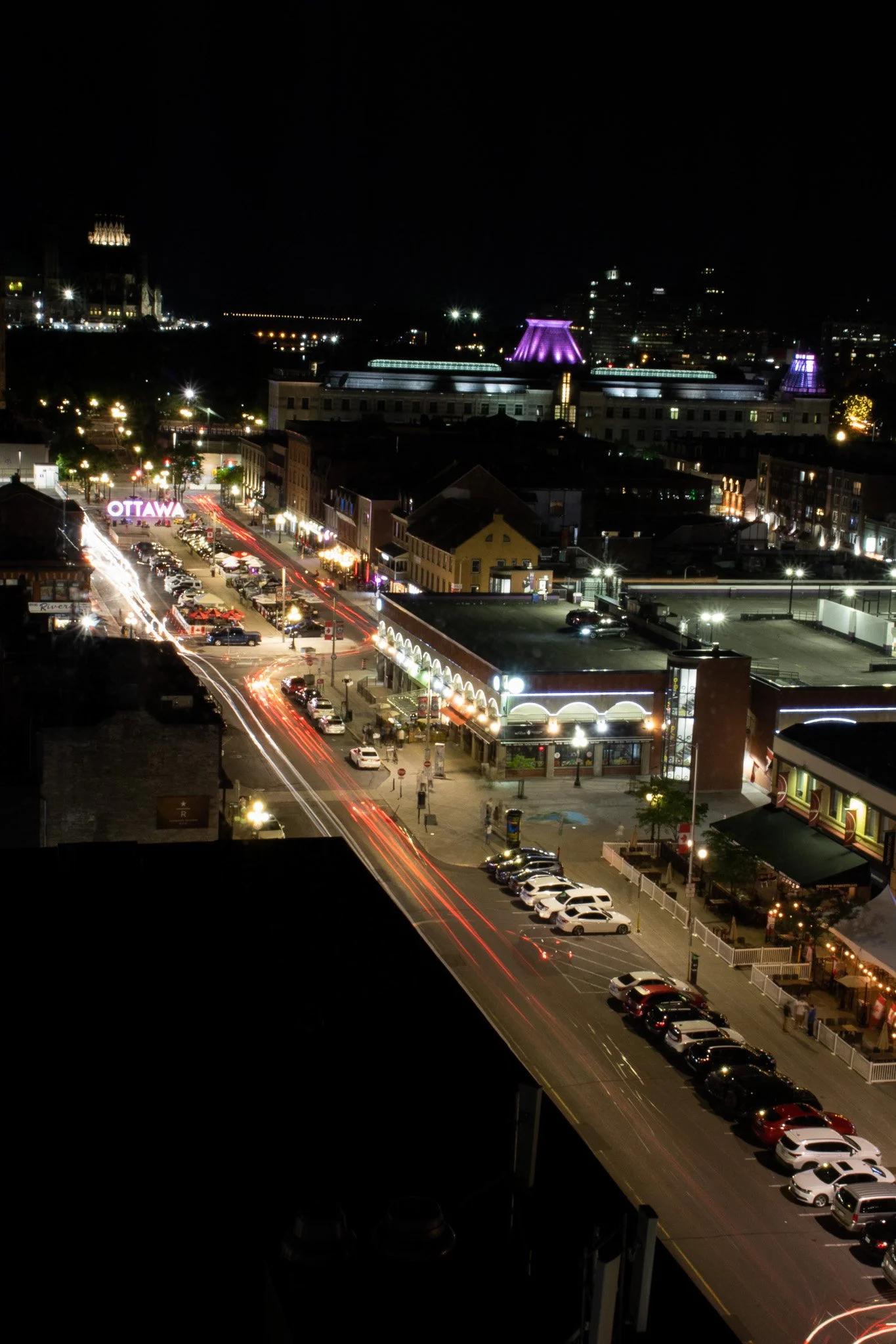 Nighttime cityscape of Ottawa with illuminated buildings, a street with light trails from moving vehicles, and several lit-up structures including a prominent sign that says 'OTTAWA'.