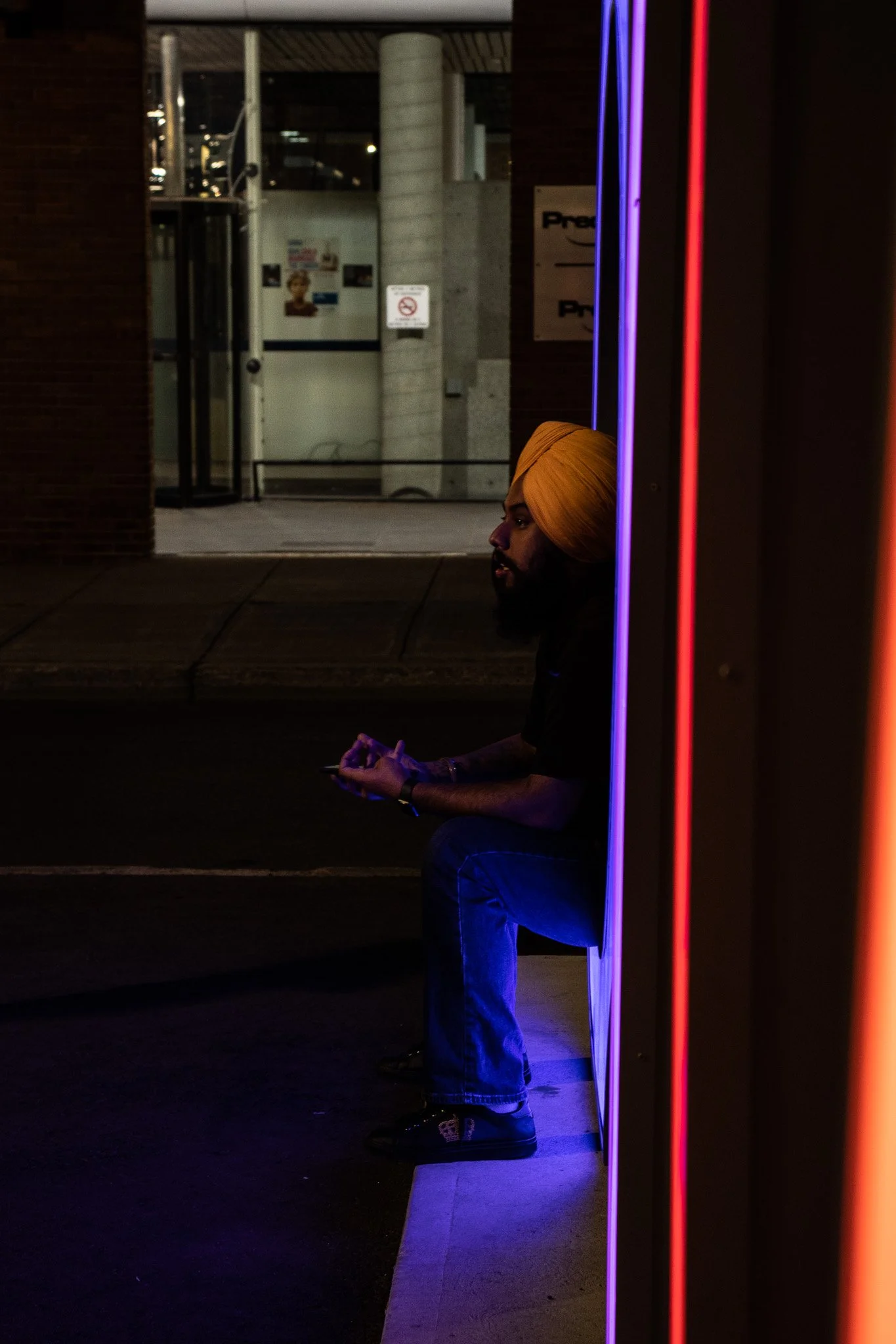 A man with a yellow turban sitting outside at night, illuminated by neon purple and red lights, holding a cellphone with a dark background of a building and some posters.