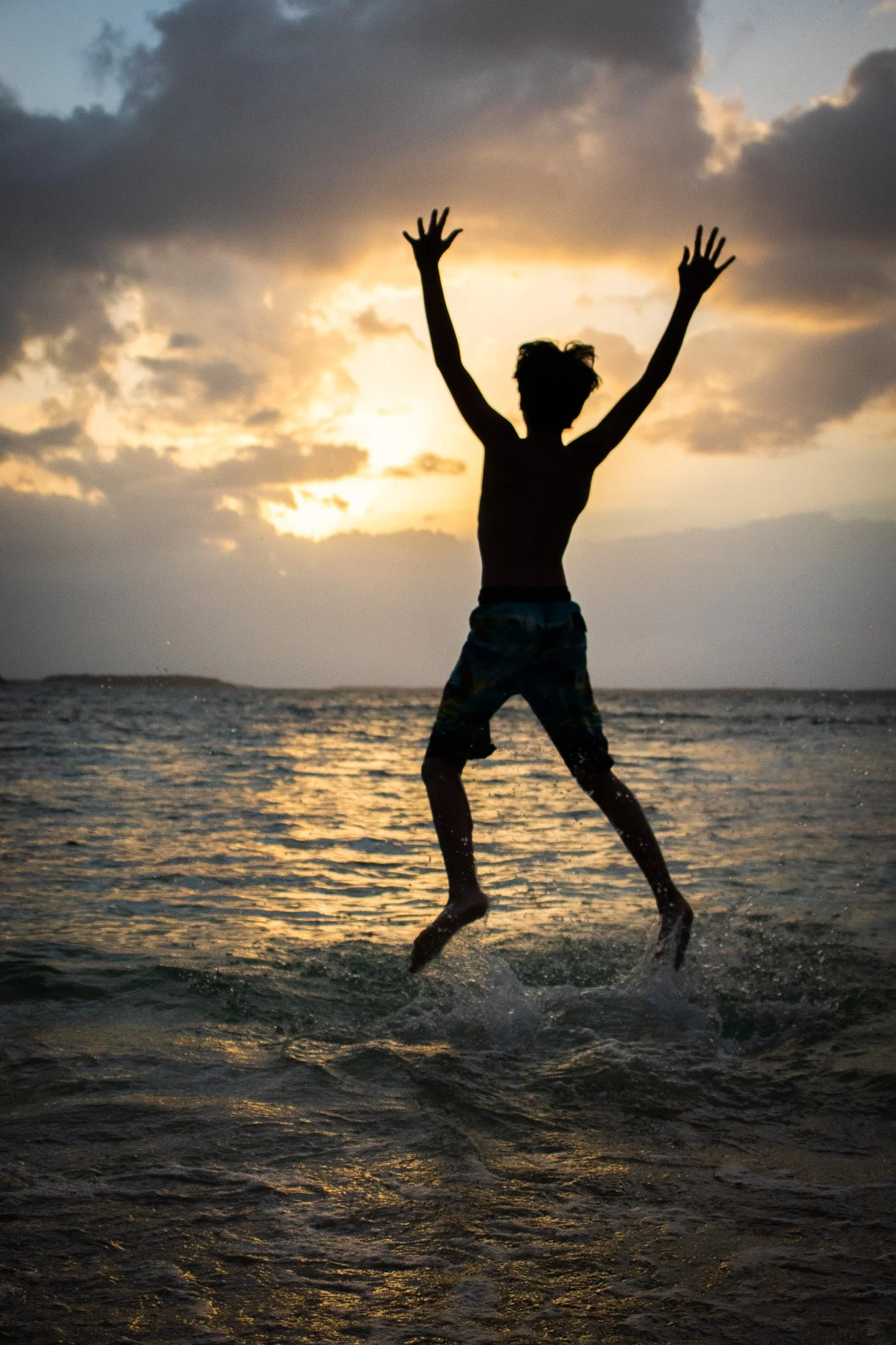 A person jumping on the beach during sunset, with arms raised above their head and wet hair, silhouetted against the sky and ocean.