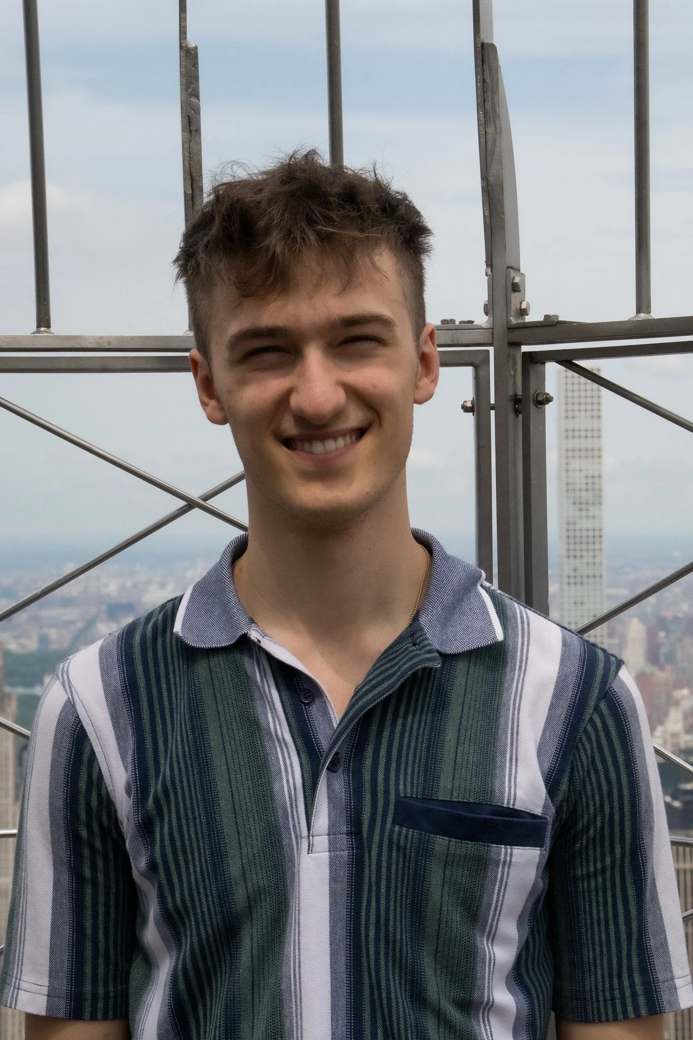 Young man with short, messy hair smiling, wearing a vertically striped polo shirt with shades of green, gray, and white, standing in front of a metal structure at a high elevation with a cityscape visible in the background.