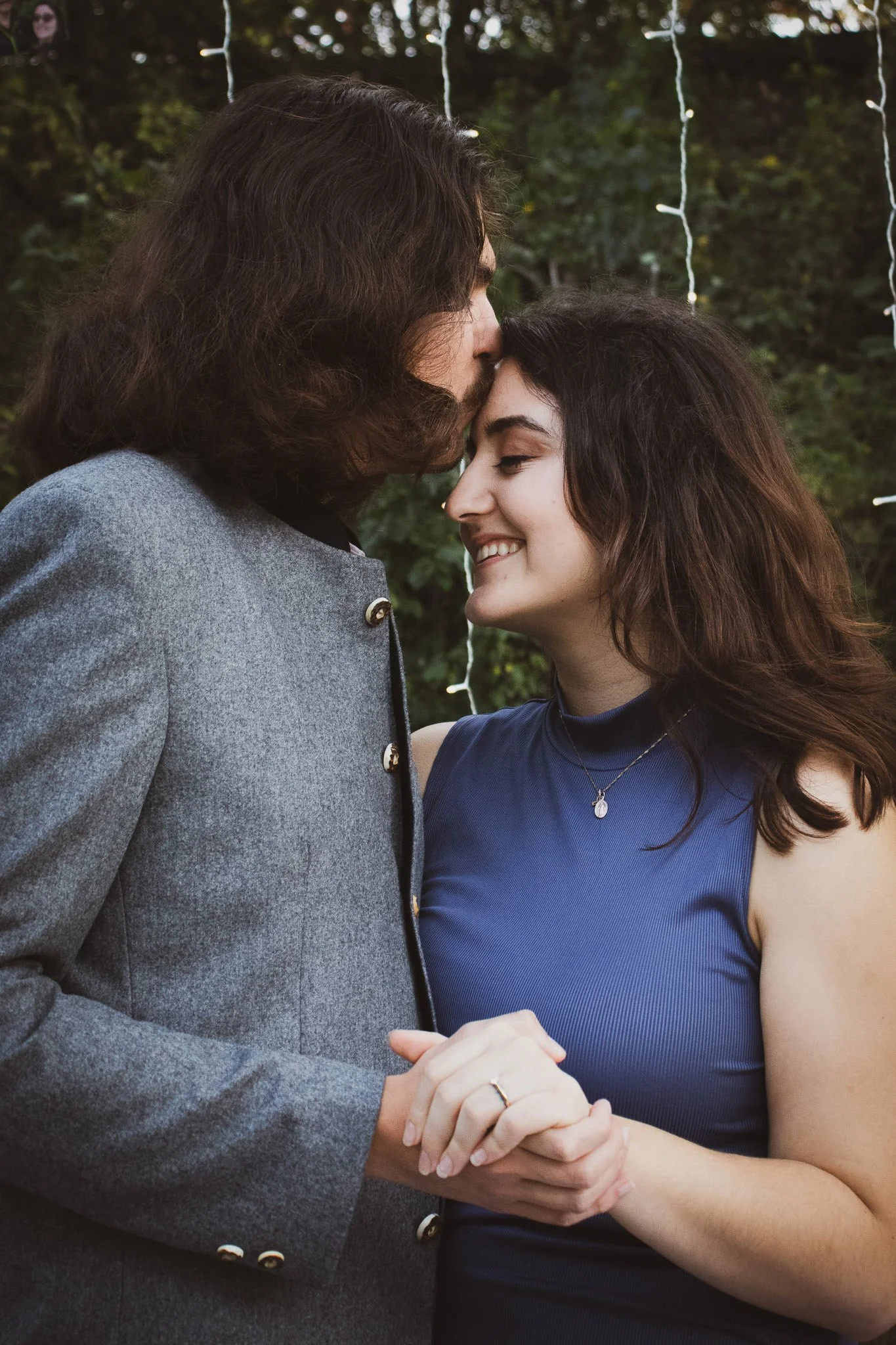 A couple is holding hands and sharing an intimate moment, with the man kissing the woman's forehead in a cozy outdoor setting decorated with string lights.
