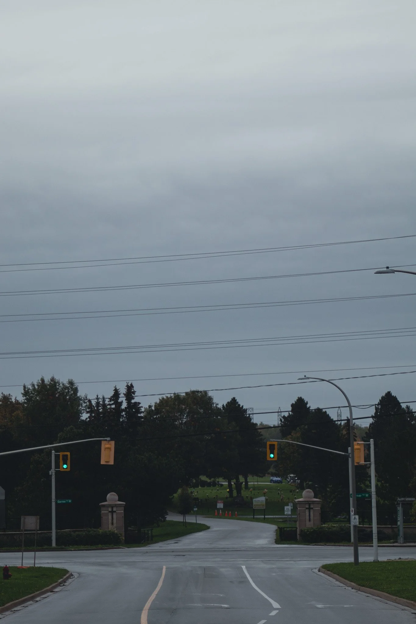 View of an empty parking lot and street with green traffic lights and street lamps, leading to a park with trees and tombstones in the distance under a cloudy sky.