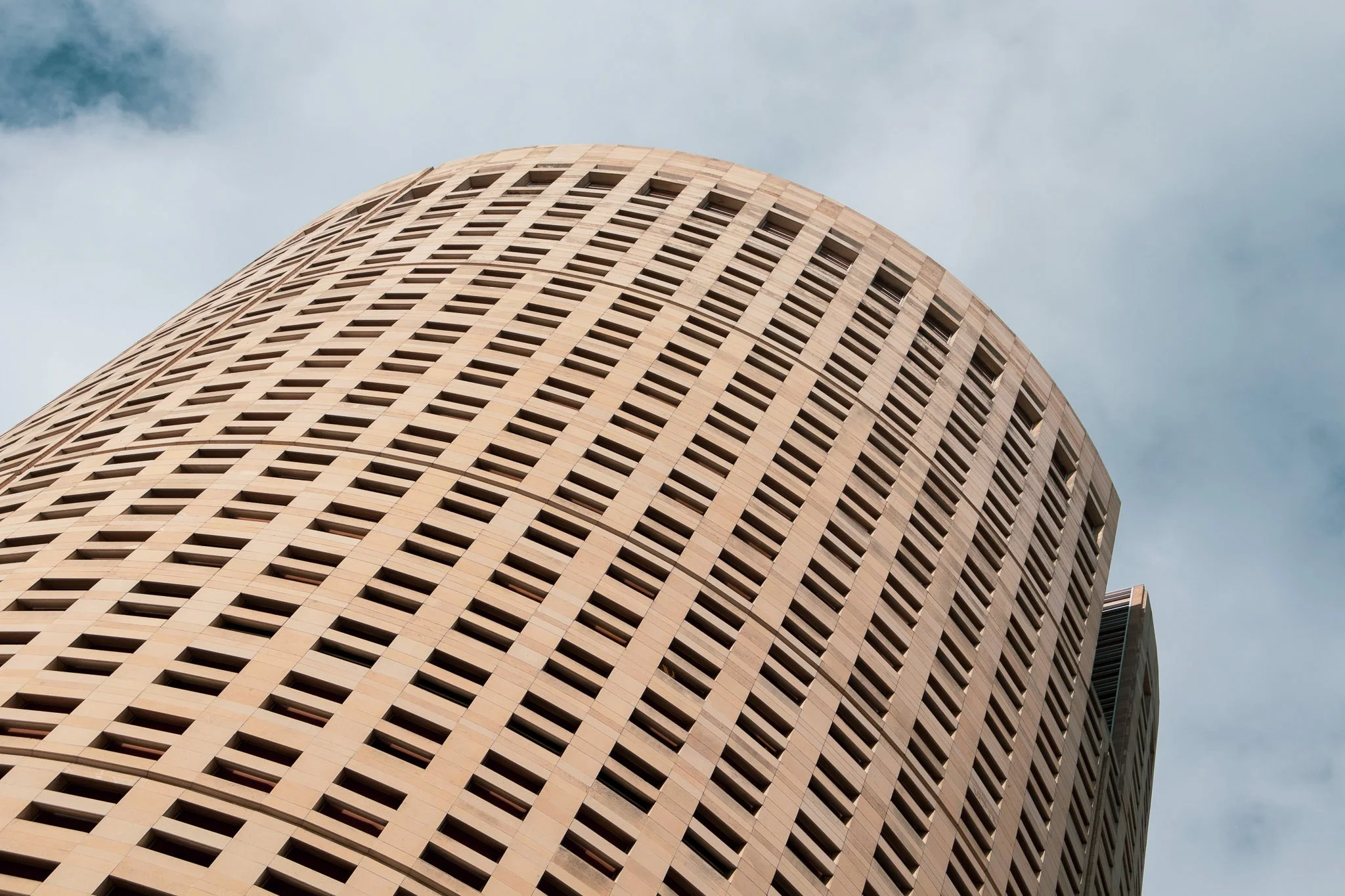 A tall, curved office building with a grid-like pattern of windows and beige exterior, viewed from below against a cloudy sky.
