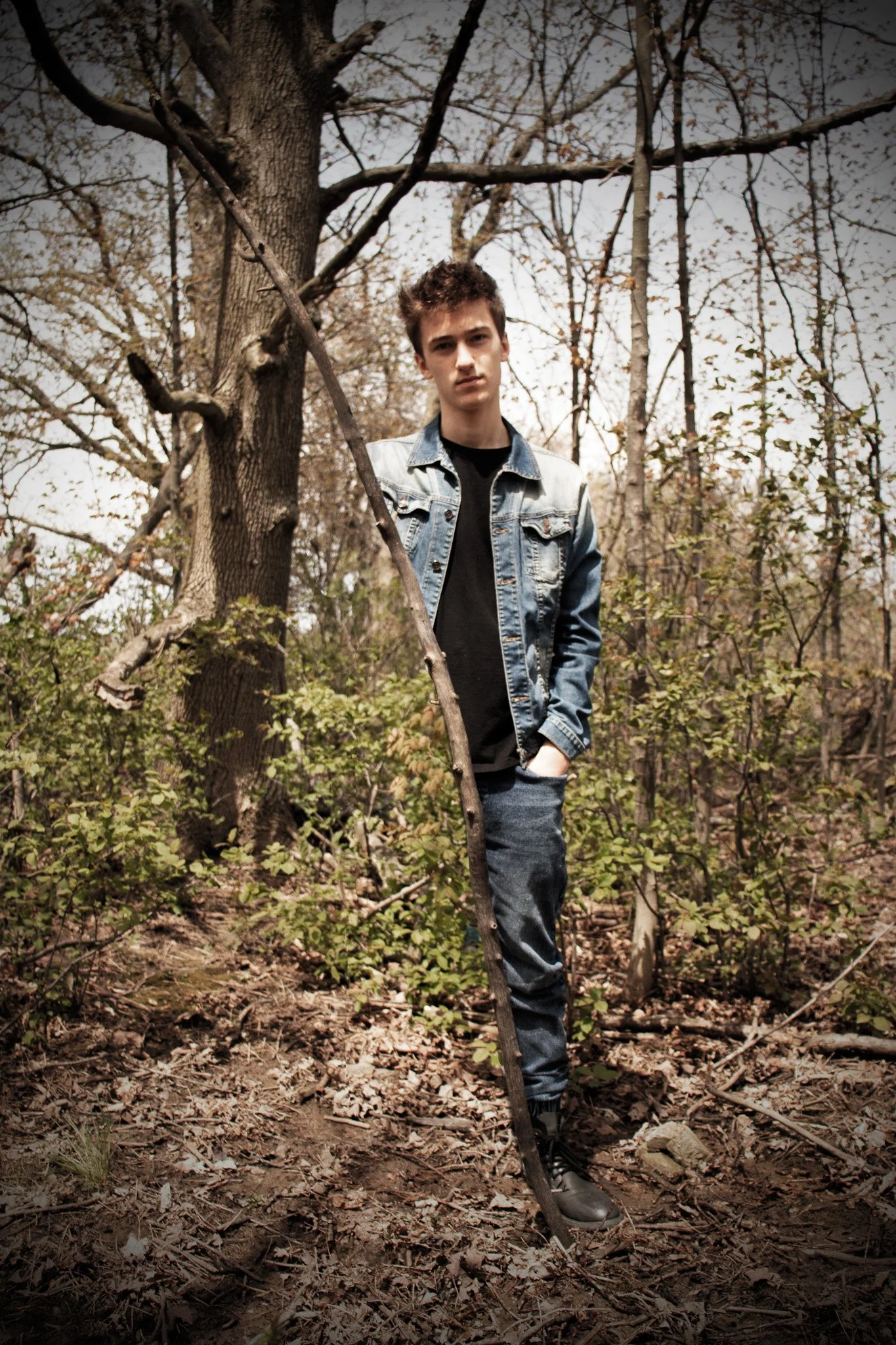 A young man stands in a wooded area, wearing a denim jacket, black shirt, and jeans, with a serious expression.