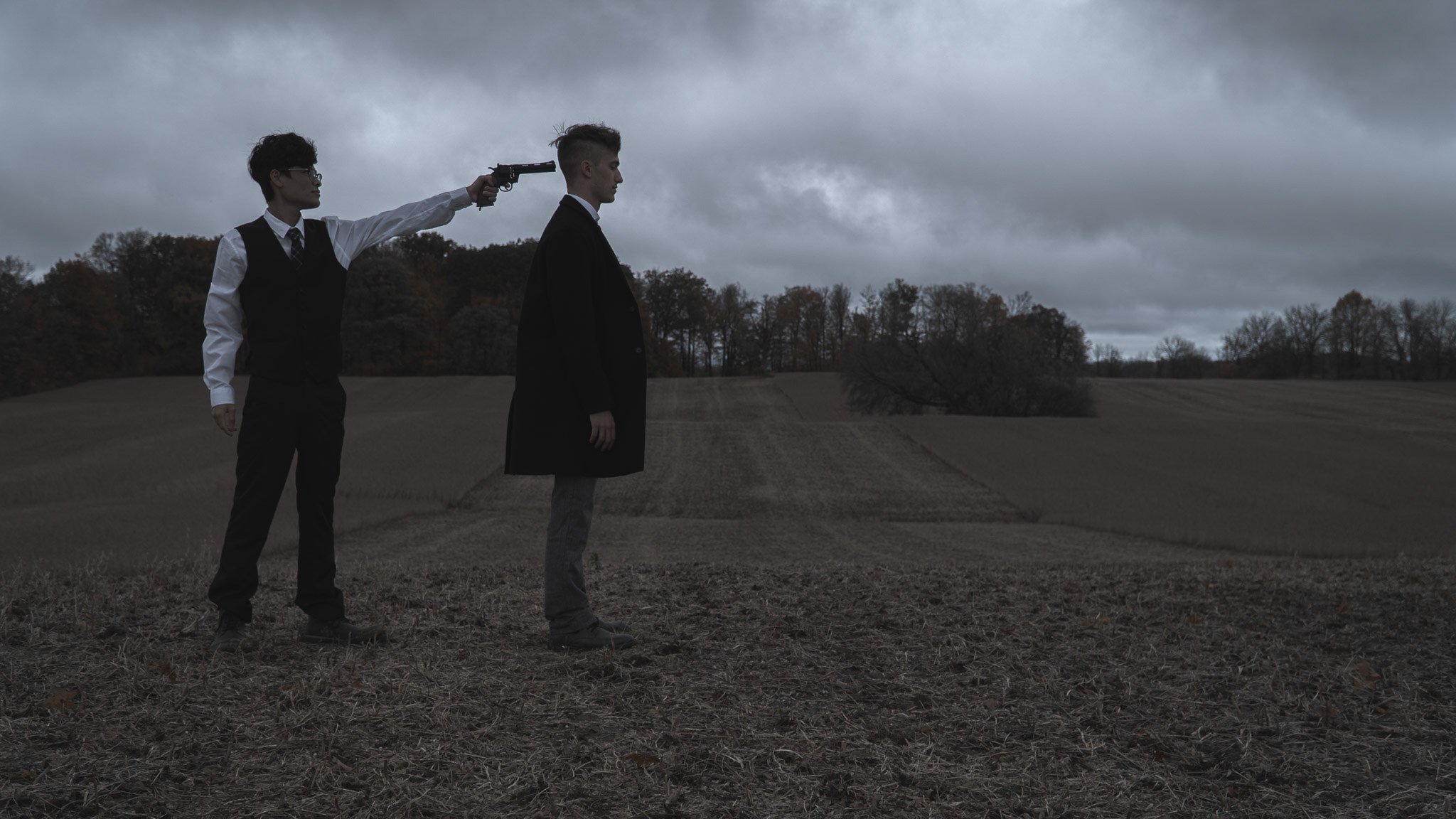 Two men in formal attire are standing in a field under a cloudy sky. One man points a gun at the other, who stands facing away.