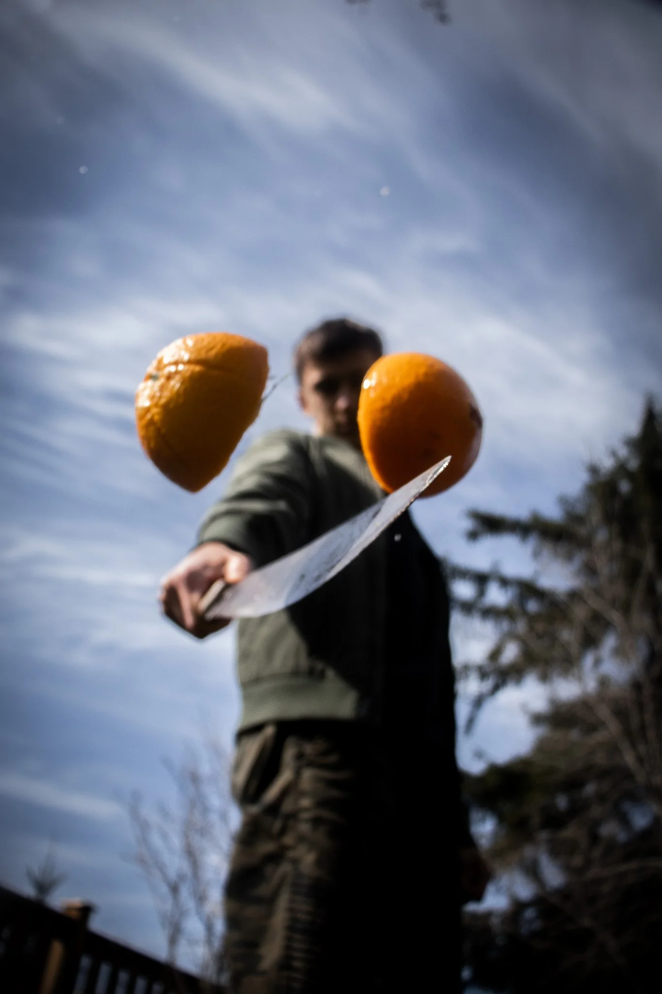 A person holding a knife towards the camera, with two oranges balanced on the knife's blade. The person is blurred in the background, and the photo is taken from a low angle with a cloudy sky overhead.