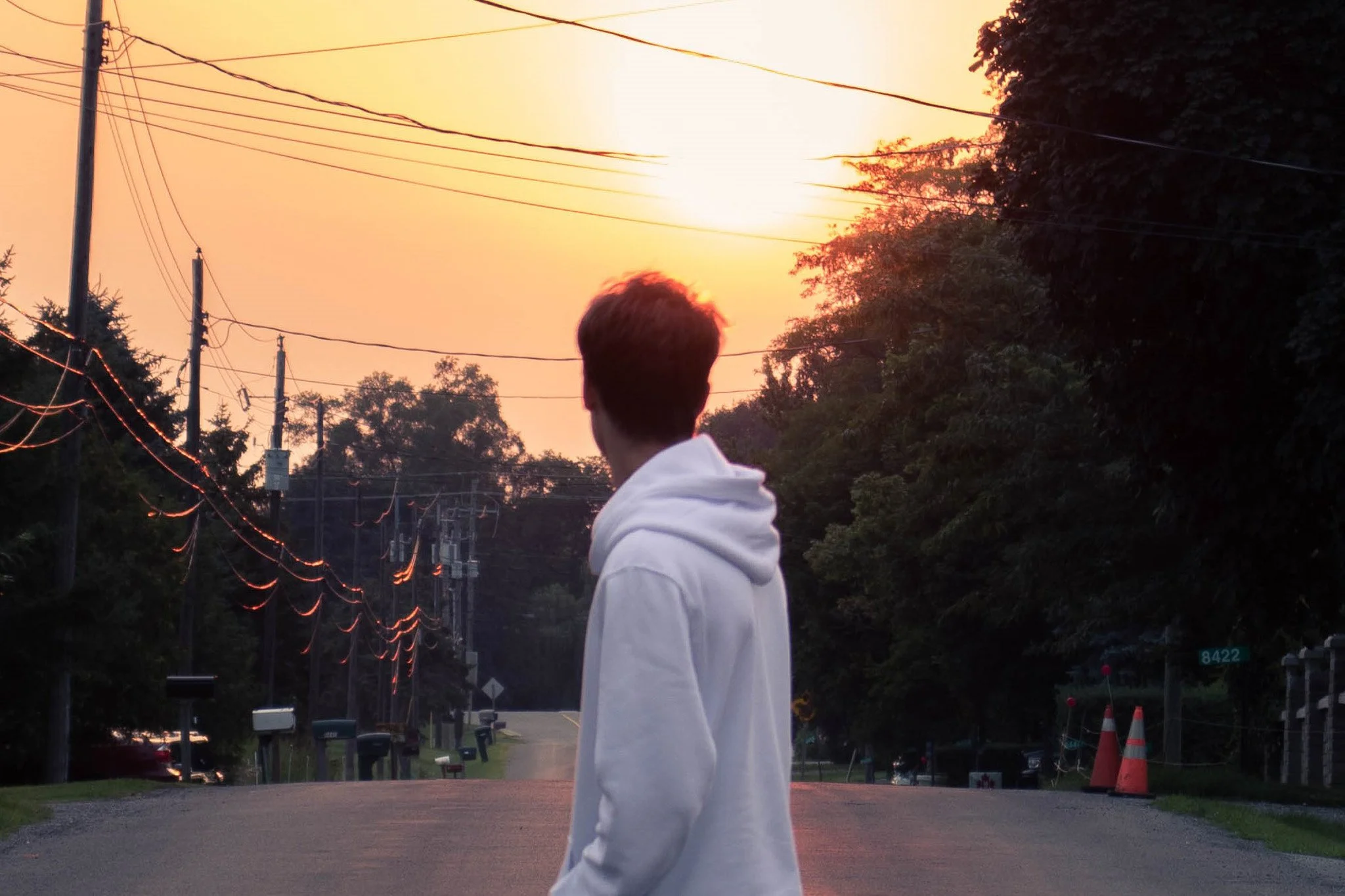 A person with short hair wearing a white hoodie standing outdoors during sunset, with power lines, trees, and traffic cones visible in the background.