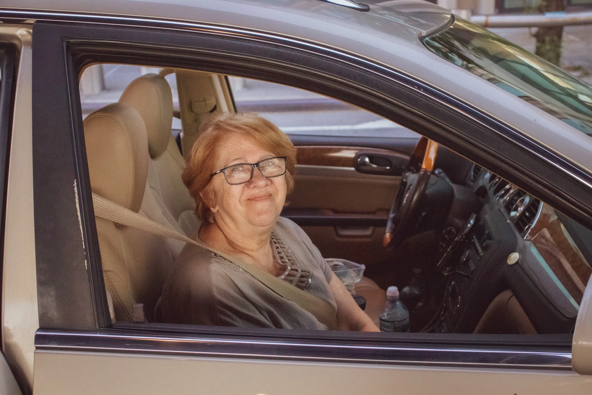 An elderly woman with glasses and short hair is sitting in the passenger seat of a car, smiling at the camera. The car has beige interior and is parked on a street.