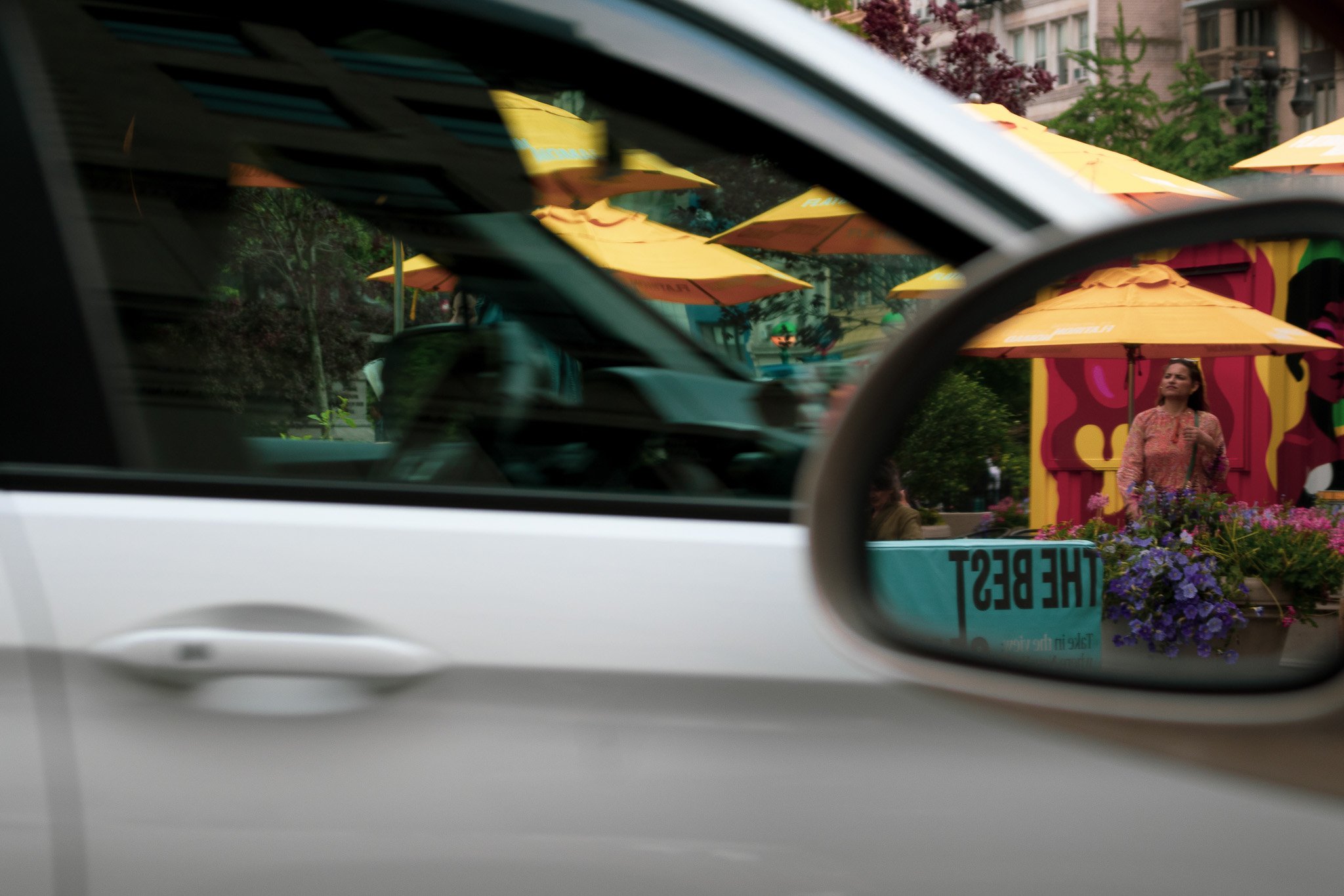 A street scene viewed through a car's side mirror showing yellow umbrellas, a woman holding flowers, and buildings.