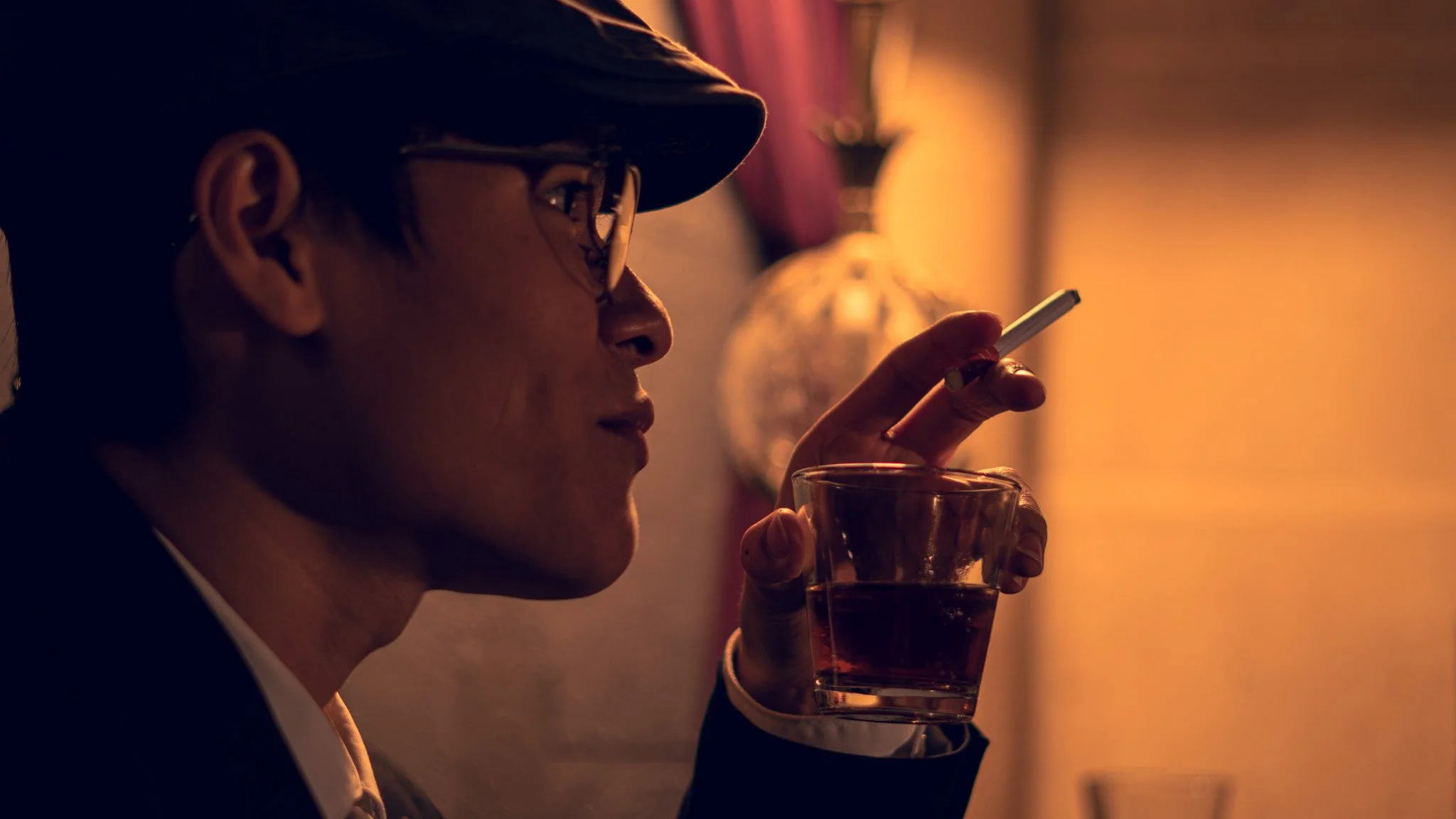 A person wearing glasses and a dark hat is holding a glass of dark beverage and looking at a smartphone in a dimly lit room with warm lighting.