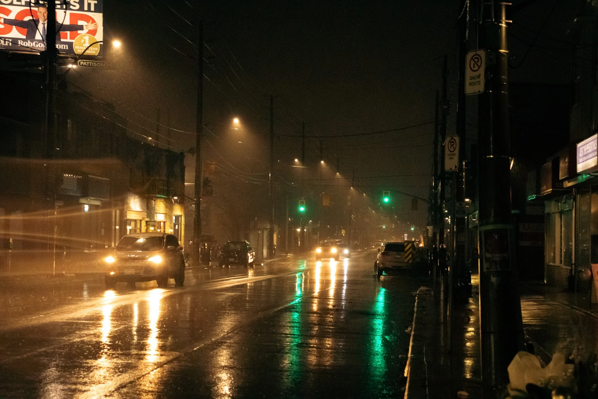 Nighttime street scene with wet pavement reflecting city lights and vehicle headlights, foggy weather, illuminated storefronts, traffic lights, utility poles, and a billboard advertising a political campaign.