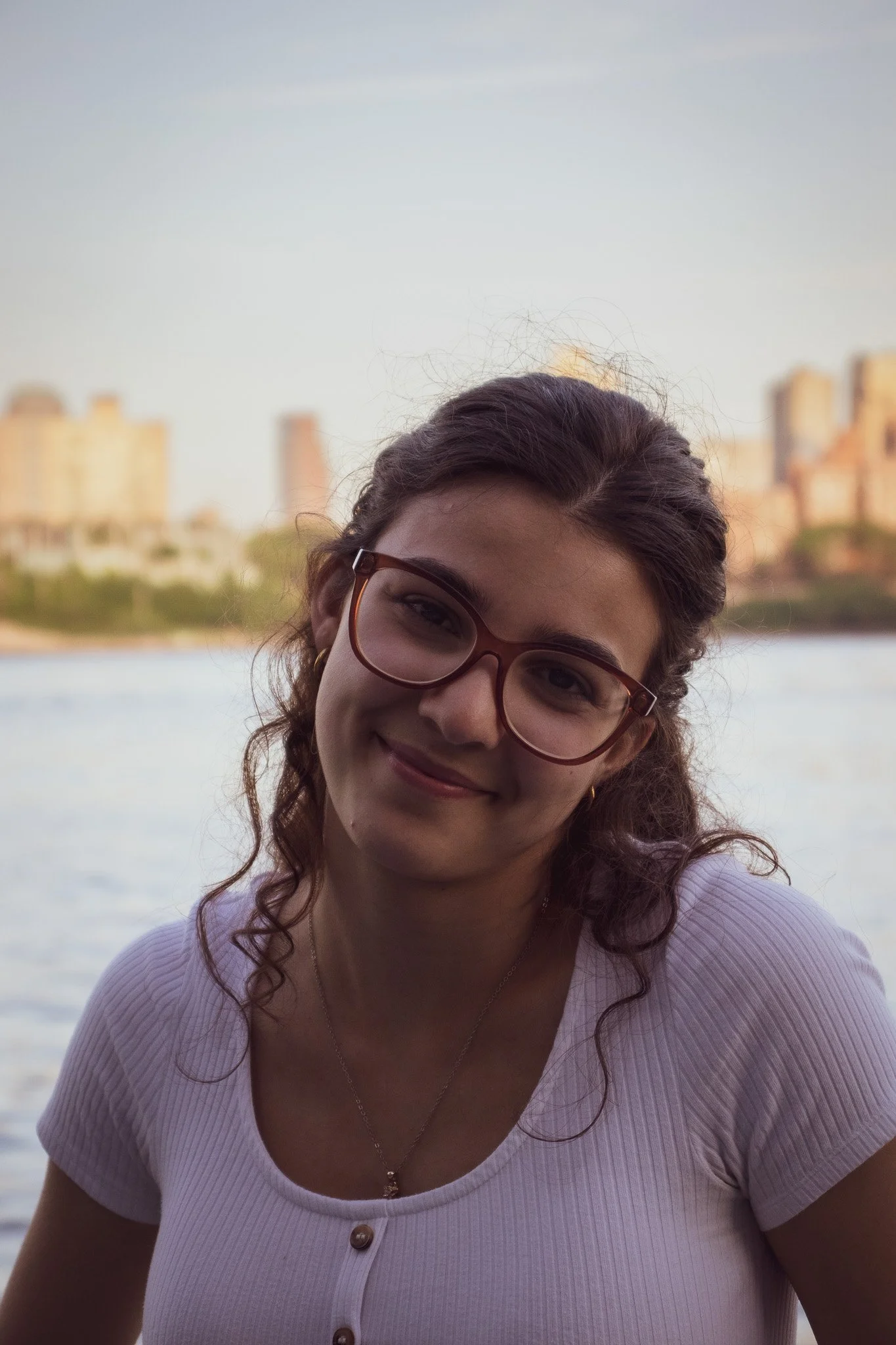 A woman with curly brown hair and wearing glasses, smiling outdoors near a body of water with a city skyline in the background.
