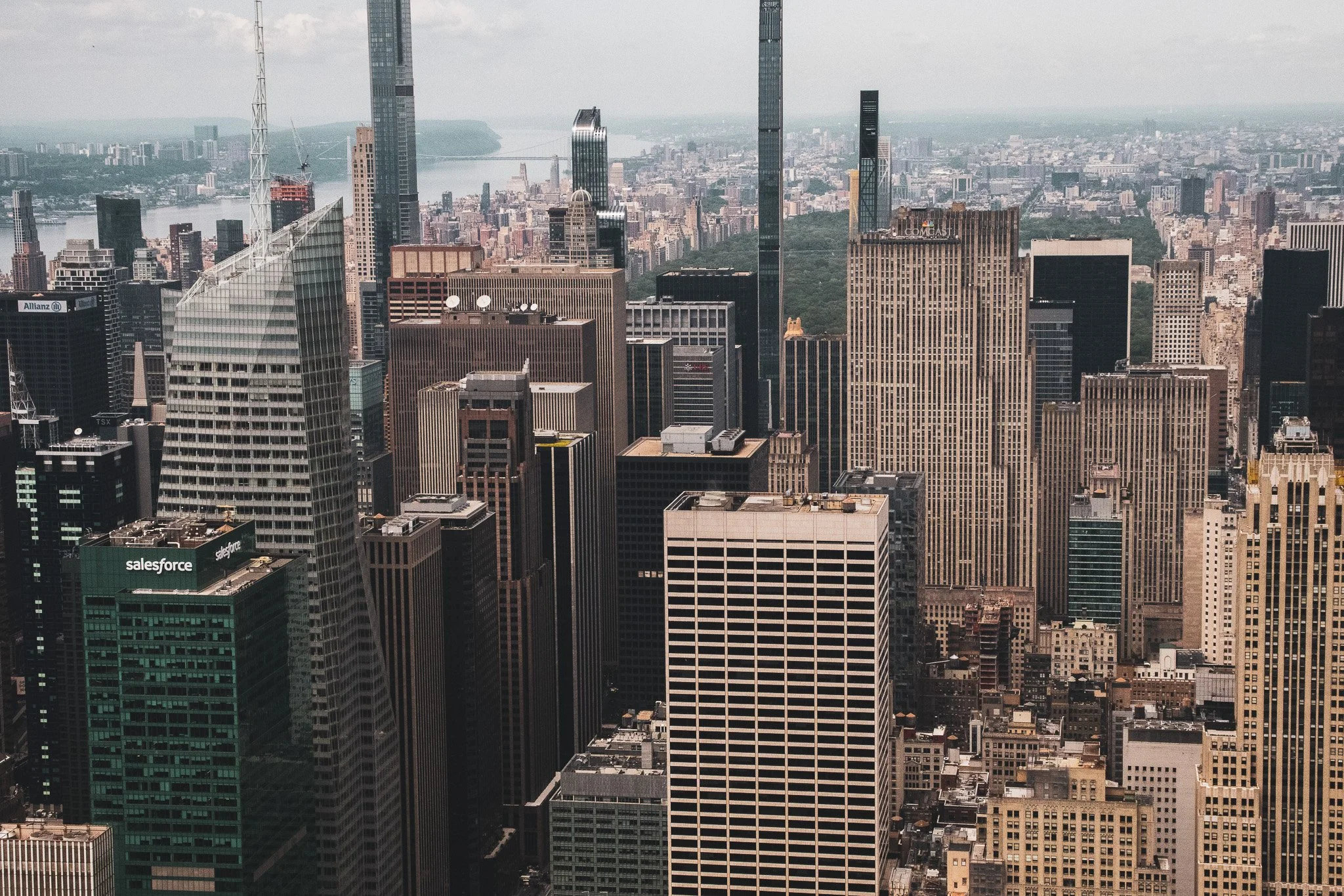 Aerial view of a dense city skyline with numerous tall skyscrapers, including buildings with signs for Salesforce and Allianz, and a river in the background.