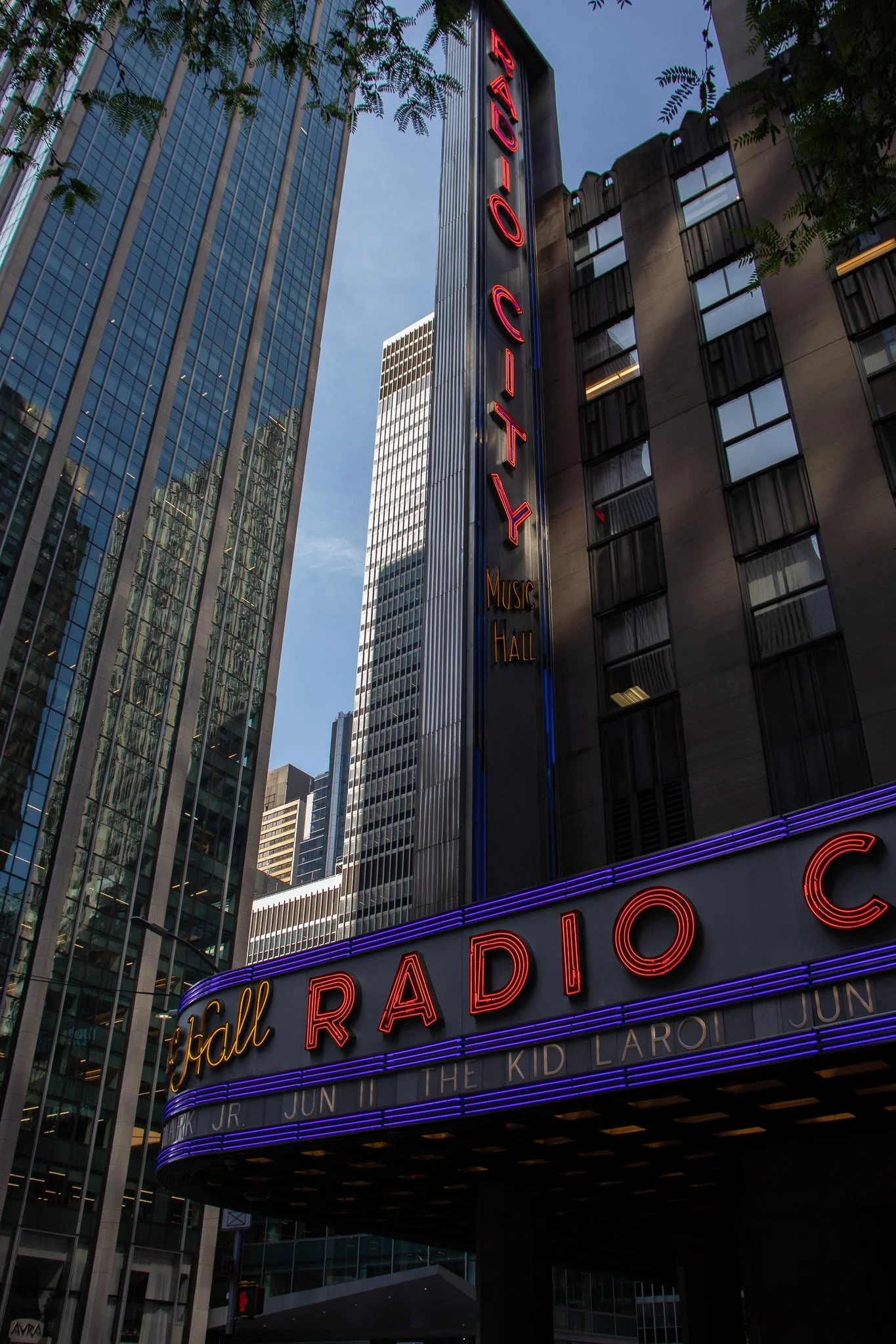 The image shows the exterior of Radio City Music Hall in New York City, with tall skyscrapers surrounding it. The marquee displays neon lights spelling out "Radio City" and other information about upcoming shows. The building's facade features large 