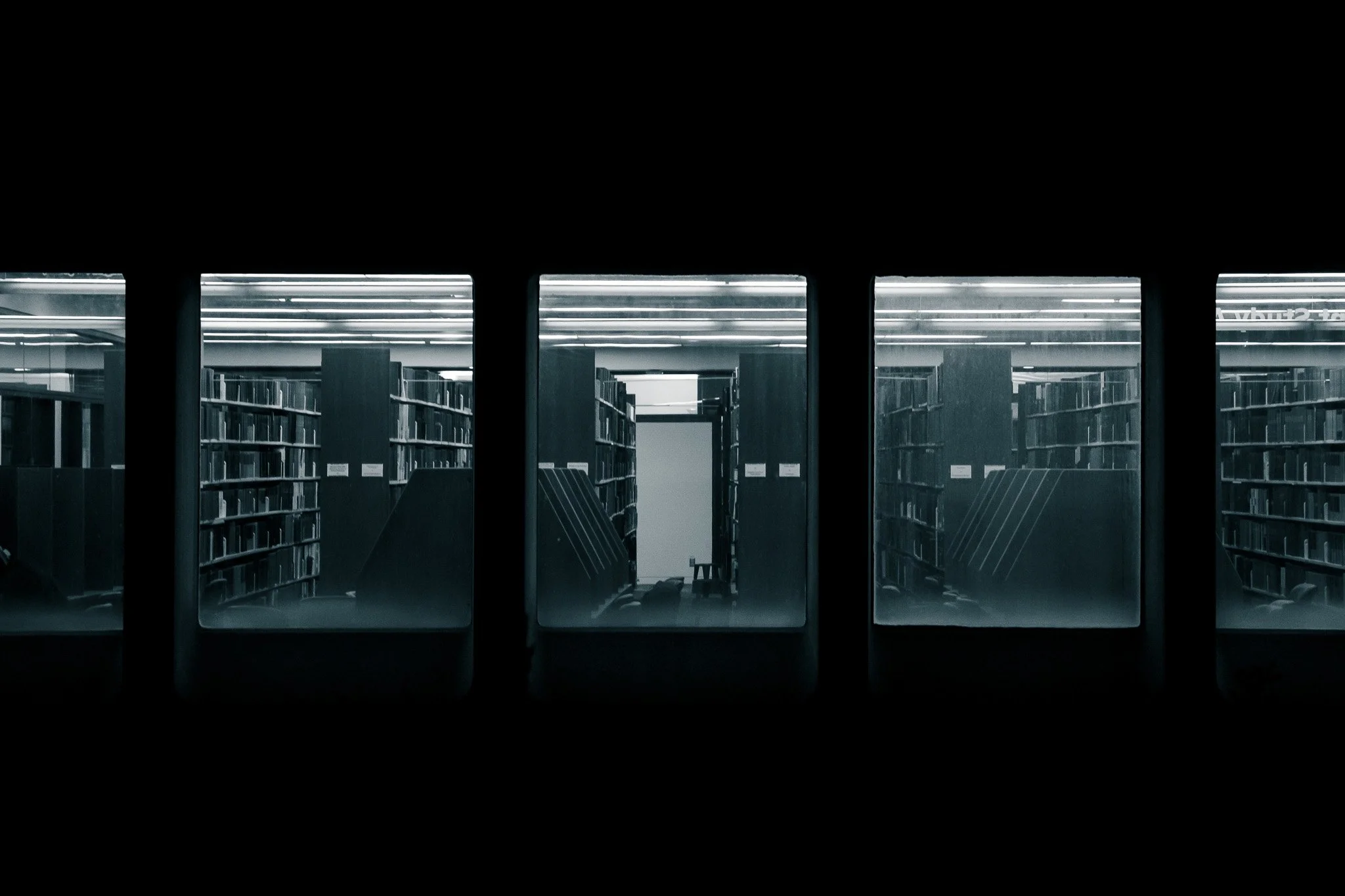 View through glass windows of a modern library interior with rows of bookshelves and fluorescent lighting.
