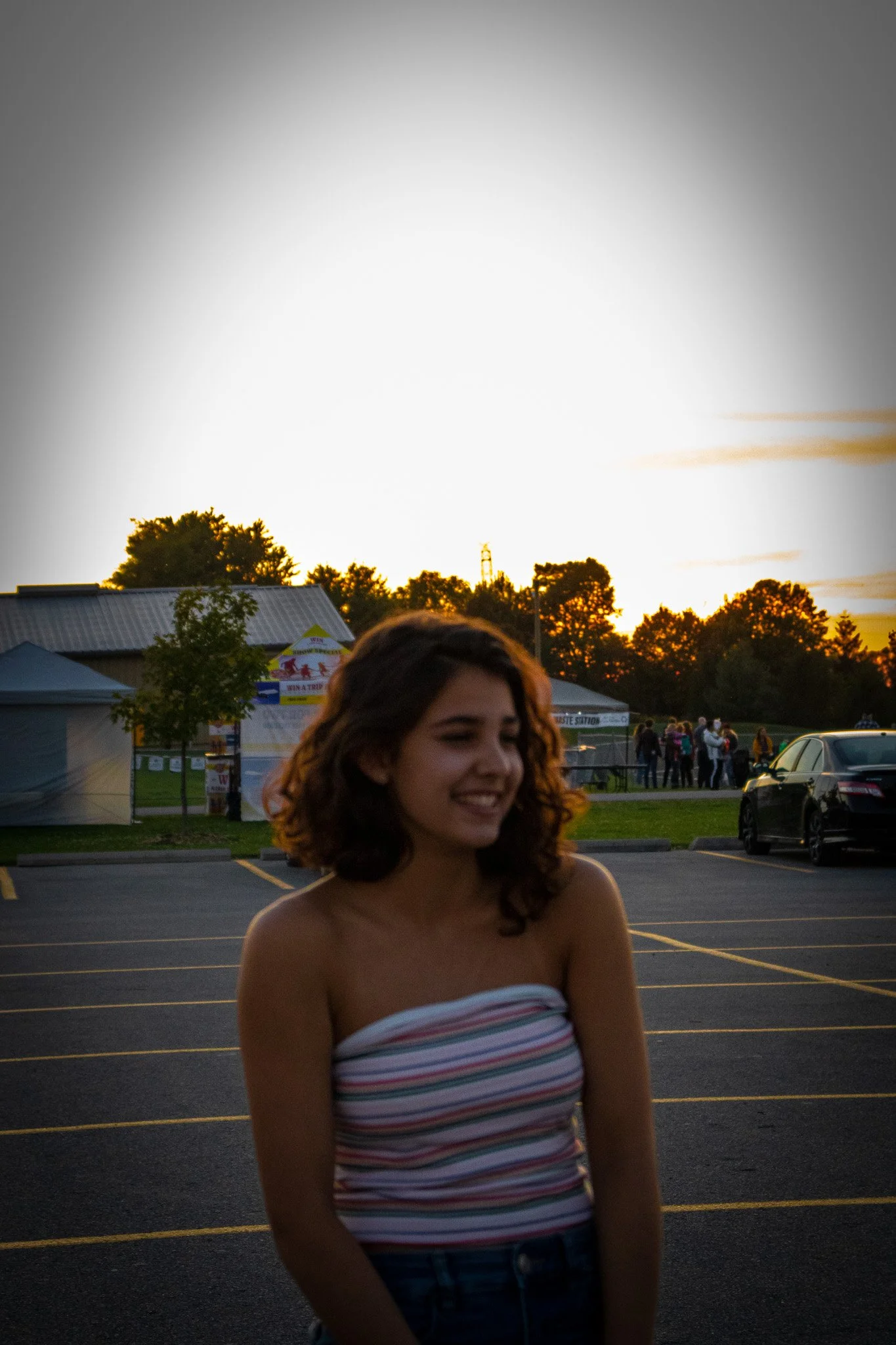 A young woman with curly hair and a strapless, striped top standing in a parking lot during sunset. There are tents and people in the background, with trees and sky visible.