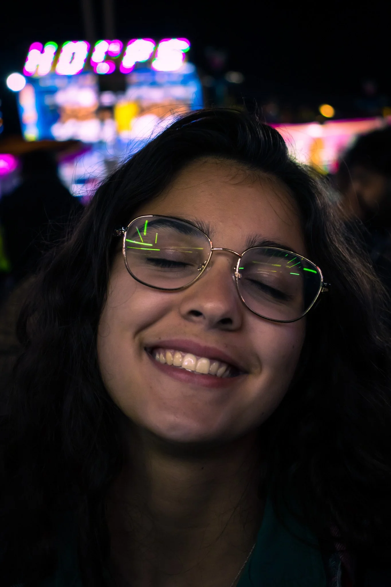 Close-up of a smiling woman with glasses, nighttime background with colorful, blurry neon lights, including 'HOPS' sign.