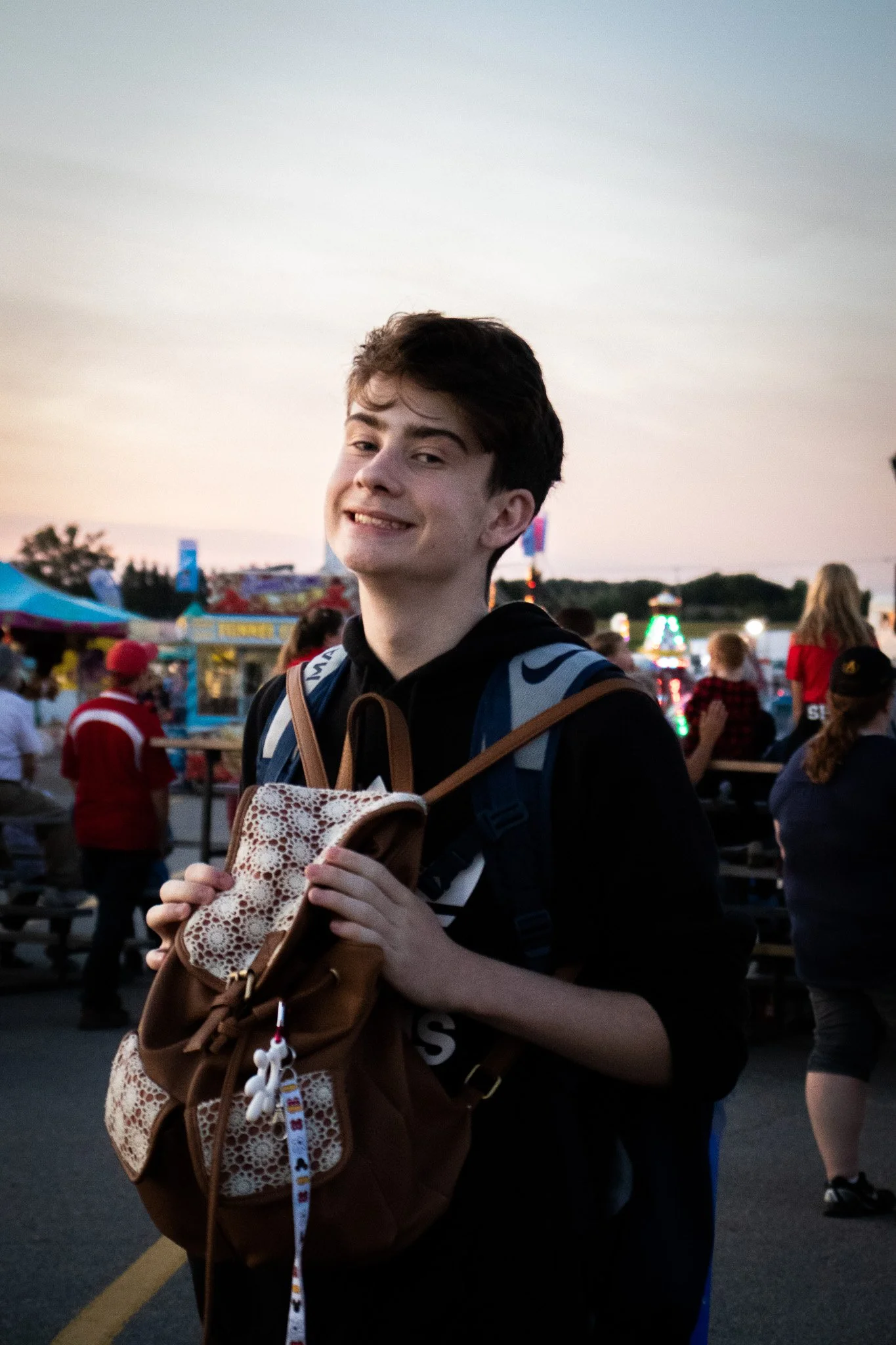 Young man smiling at a fair or carnival, holding a small purse with a teddy bear keychain, with rides and booths in the background during dusk.