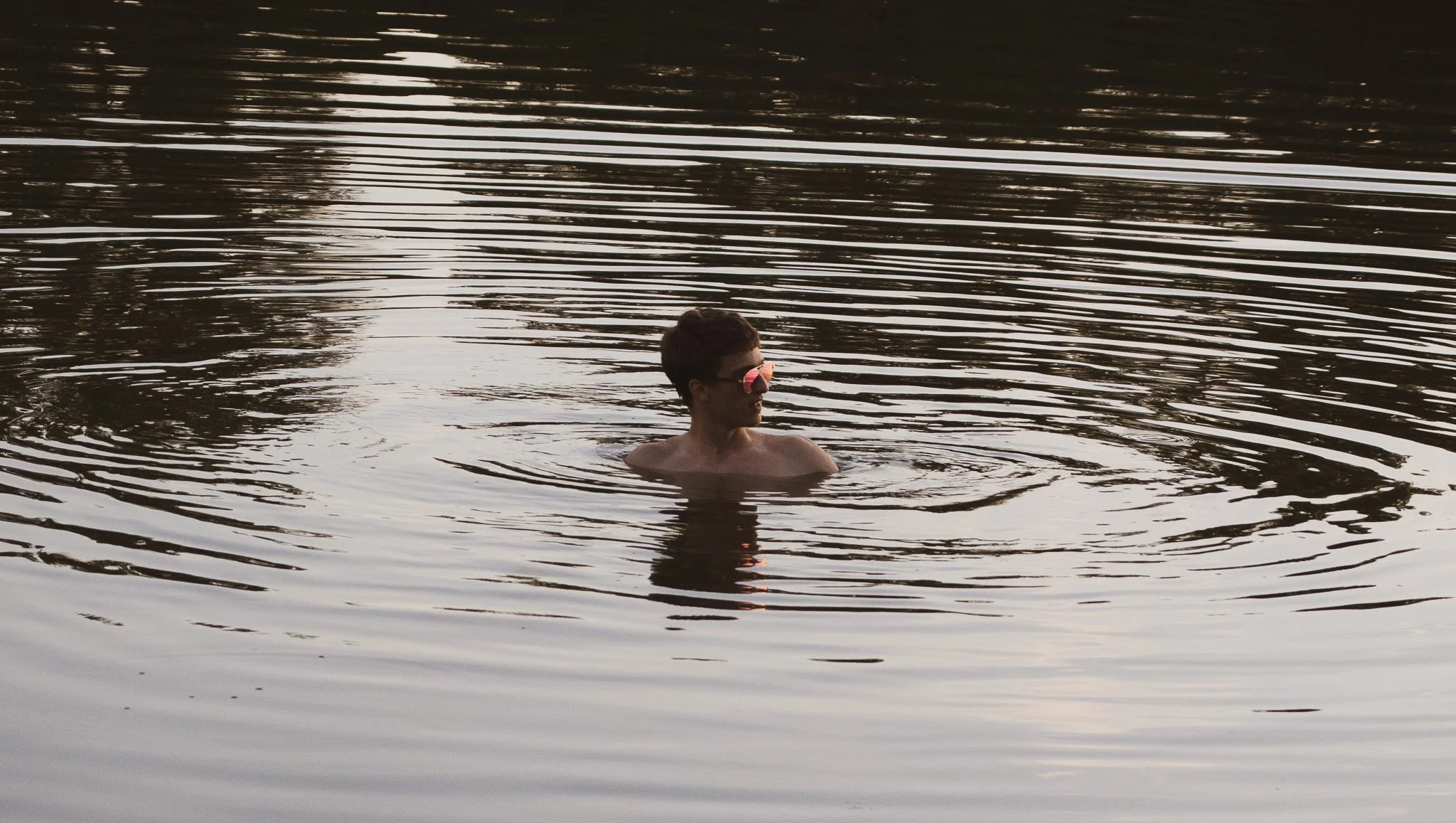 A young man with sunglasses swimming in a calm body of water during sunset.