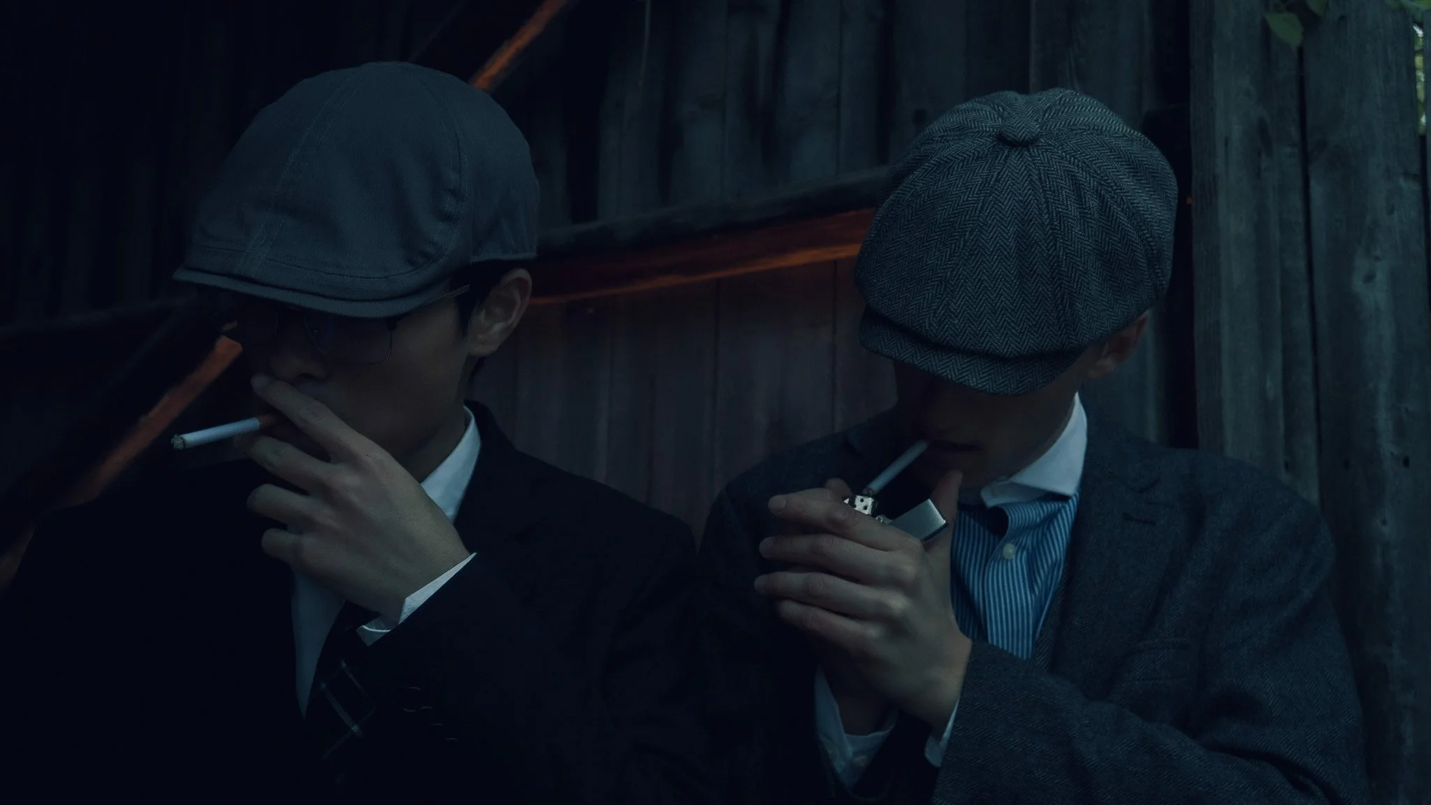 Two men wearing suits, ties, and hats smoking cigarettes in a dimly lit wooden setting.